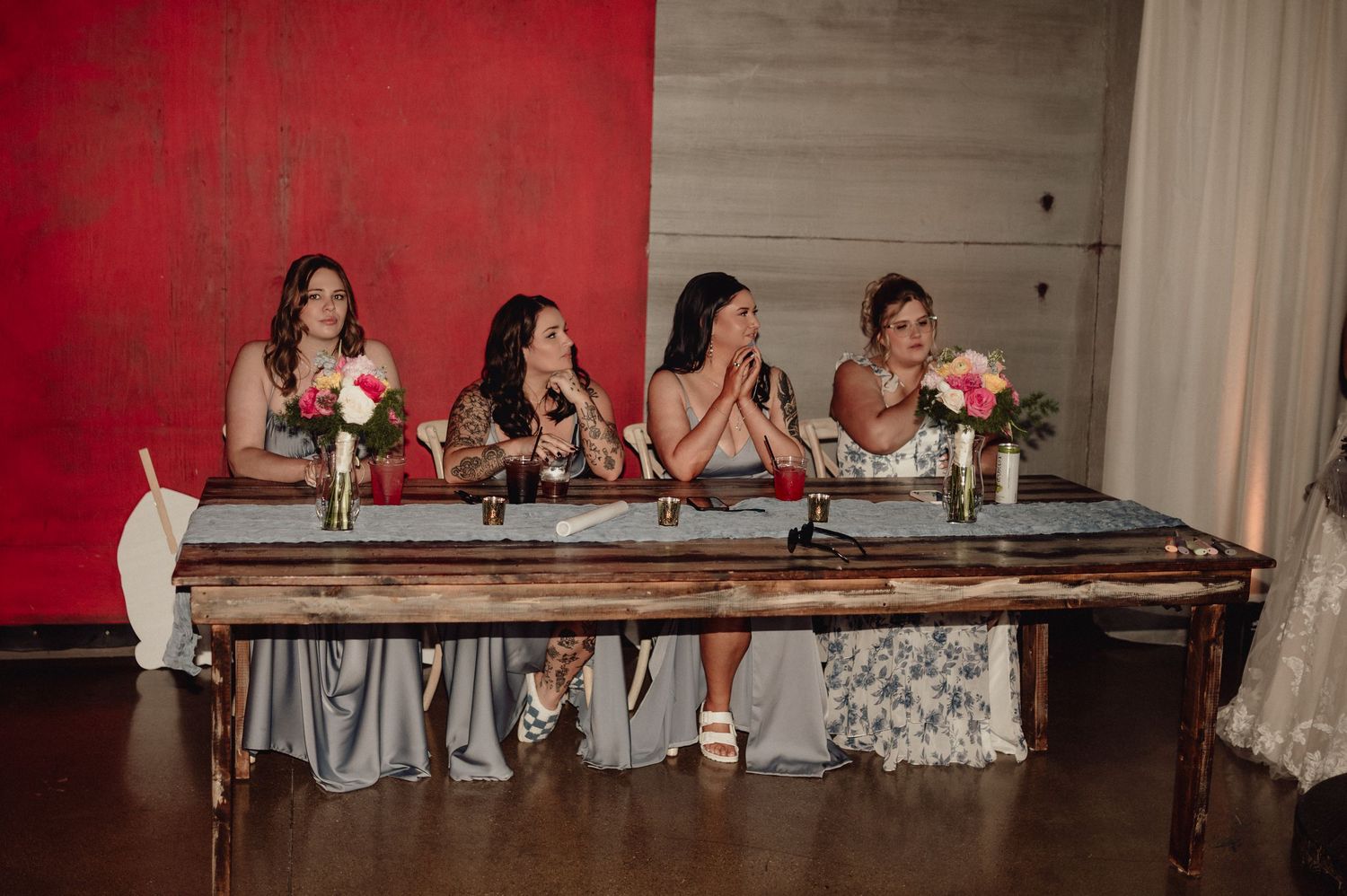 Wedding party members sit at a long table against a red wall during a reception celebration.
