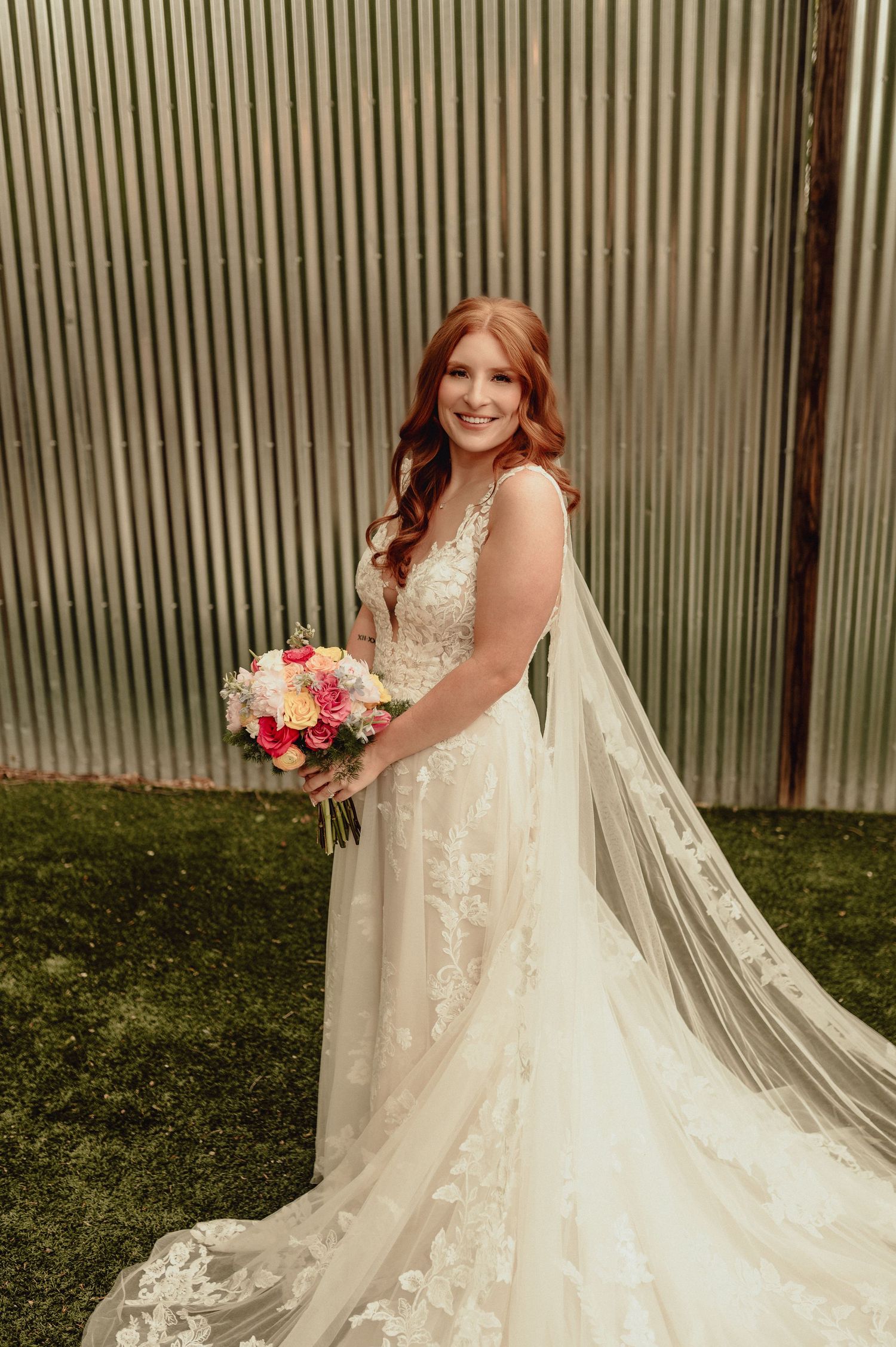 Bride in a flowing lace wedding gown with cathedral veil poses in front of corrugated metal wall with grass.