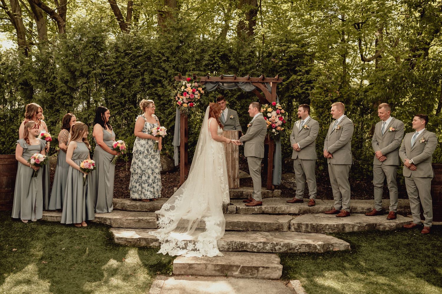 Wedding party stands in a garden setting during an outdoor ceremony with stone steps and lush greenery.