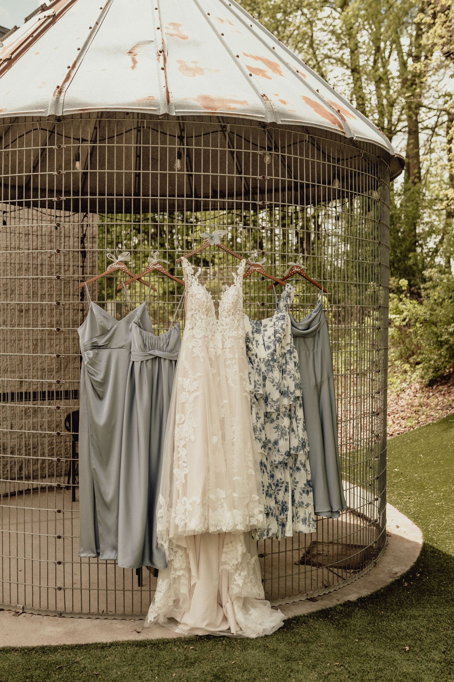 Wedding dresses and bridesmaid gowns hanging from vintage iron gazebo in outdoor garden setting.