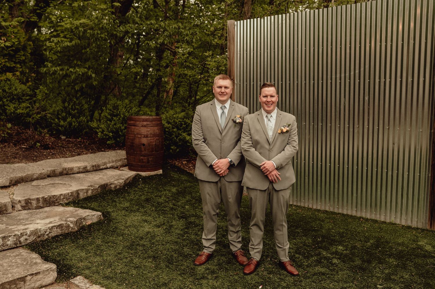 Two people in light suits sit on a stone wall in front of a white fence and garden backdrop.