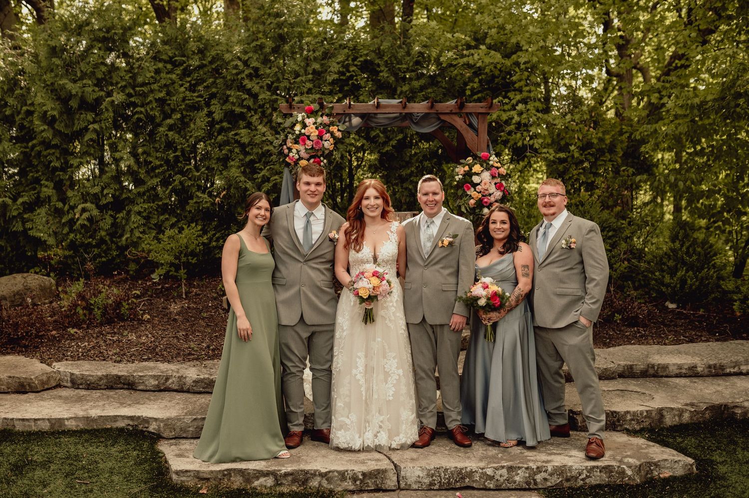 Wedding party in sage green and ivory attire poses together outdoors during golden hour in a garden setting.