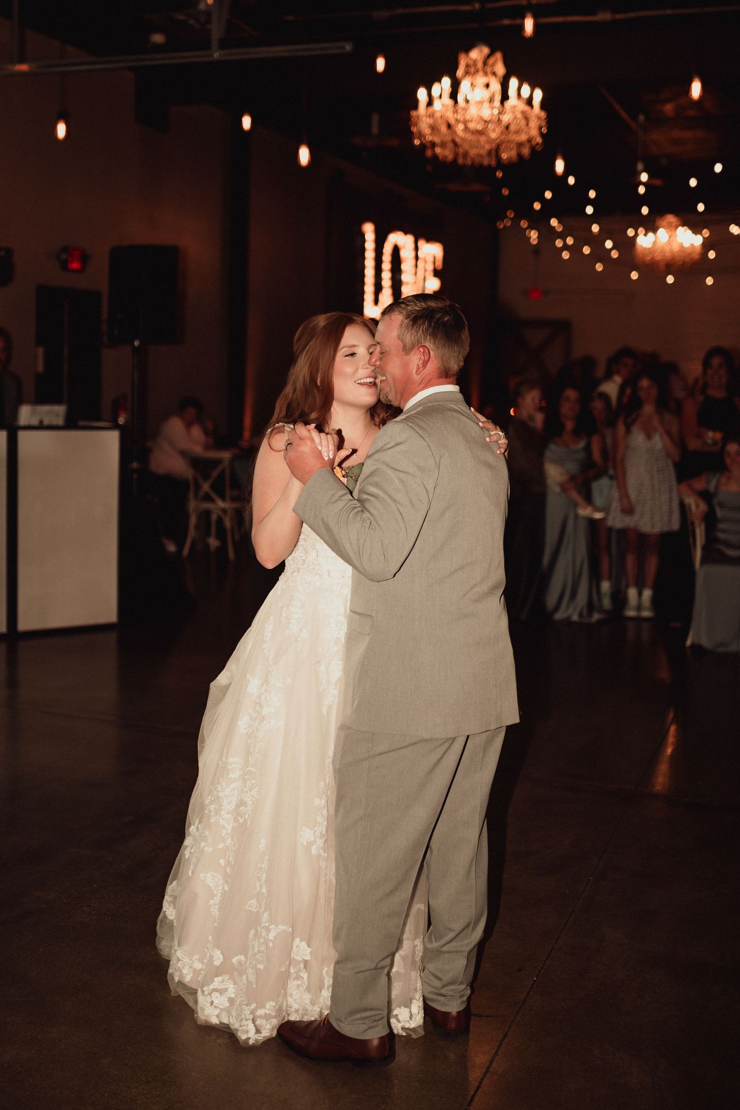 A romantic wedding couple shares a dance together under glowing chandeliers in a dimly lit ballroom.