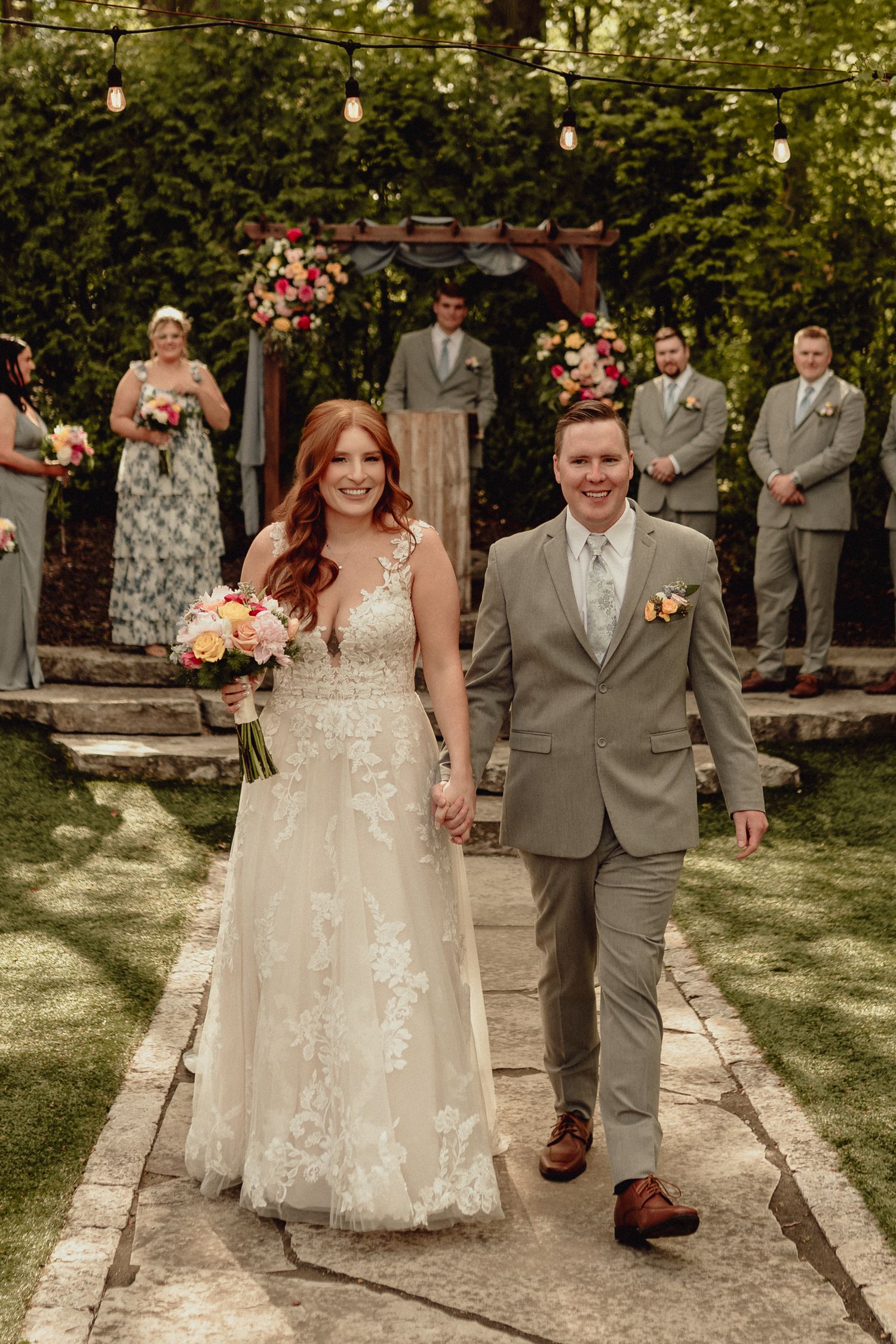 Wedding party walks along stone pathway in garden setting with string lights overhead and lush greenery in background.
