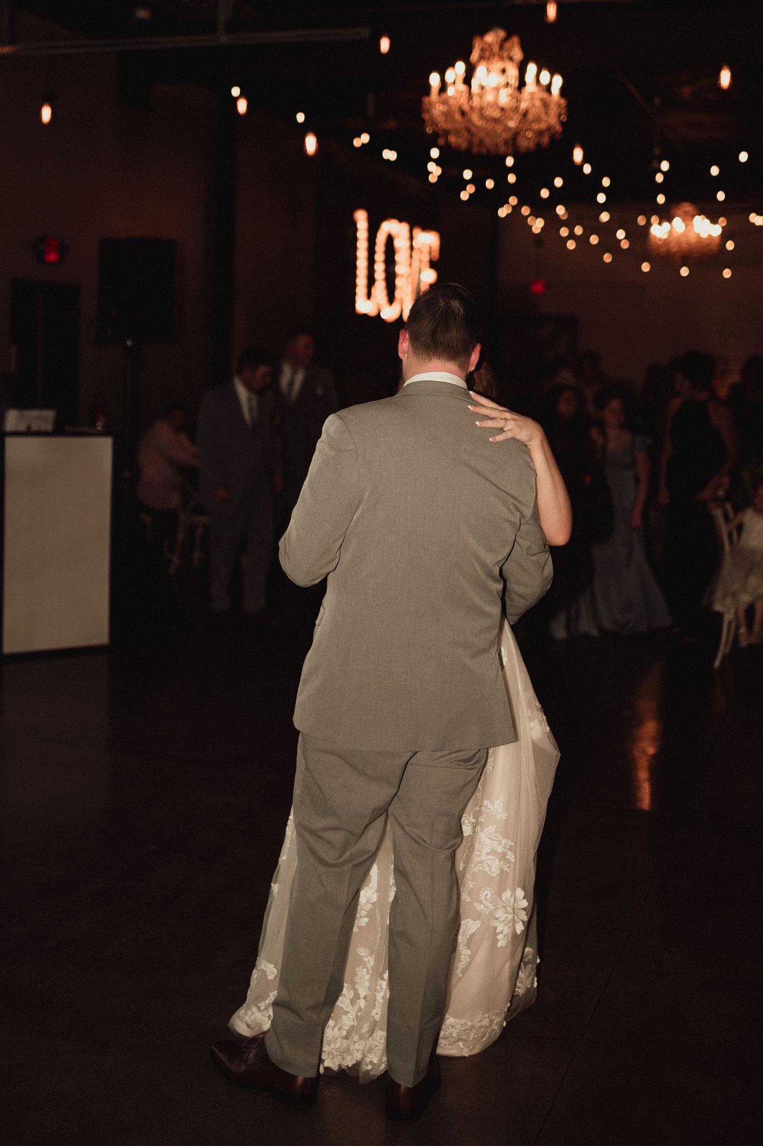 A bride and groom dance together on a dimly lit dance floor under twinkling lights and chandeliers at their wedding reception.