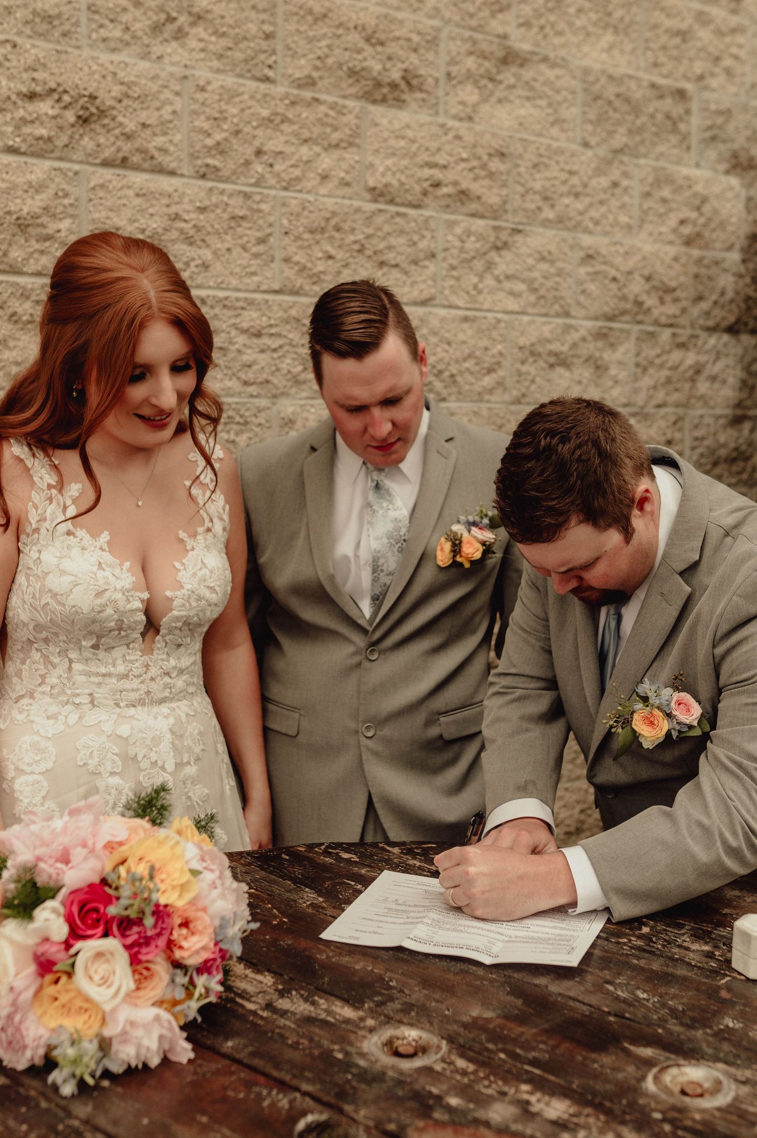Bride and groom sign marriage documents together at rustic wooden table with colorful floral bouquet.