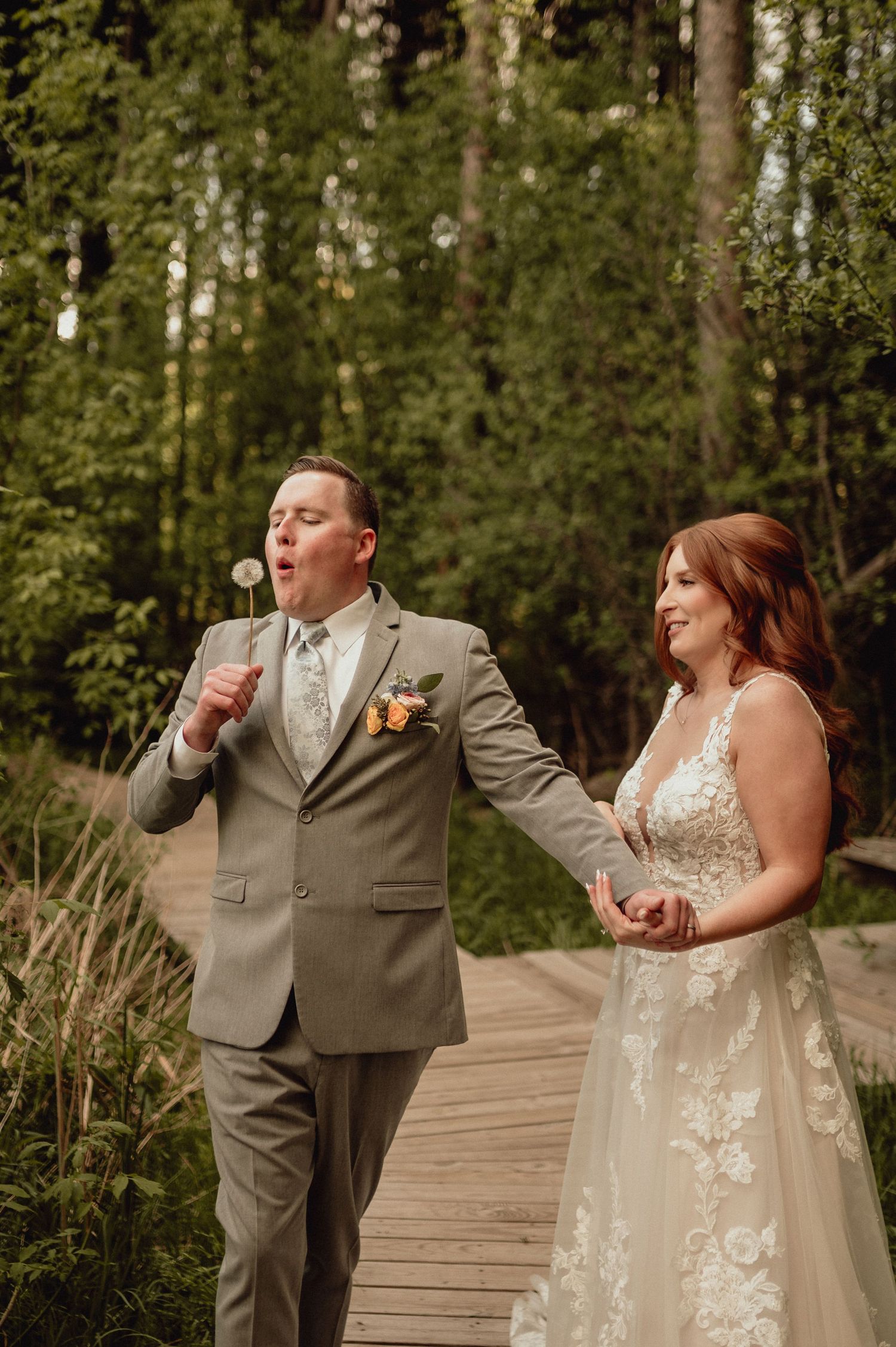 A couple in wedding attire share a candid laughing moment together on a wooden boardwalk surrounded by trees.