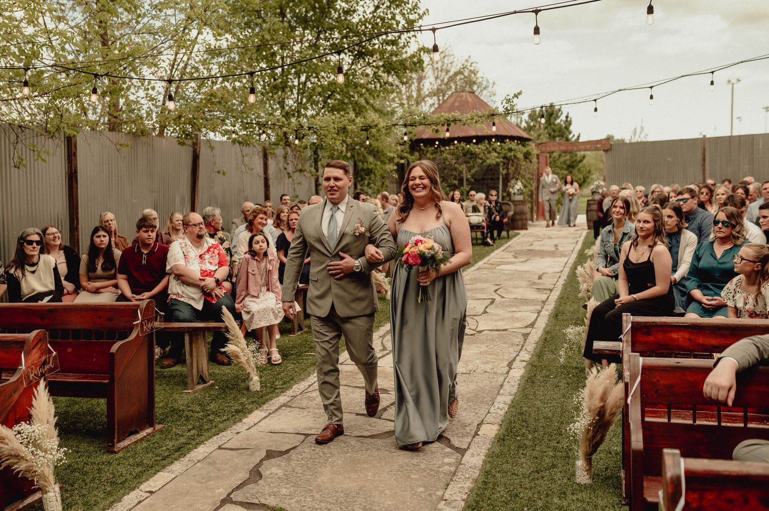 Wedding guests sit on rustic wooden benches during an outdoor ceremony with string lights overhead.