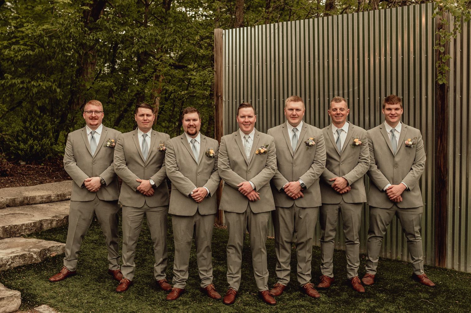 A wedding party of seven groomsmen wearing matching gray suits and brown shoes pose in front of a metal wall outdoors.