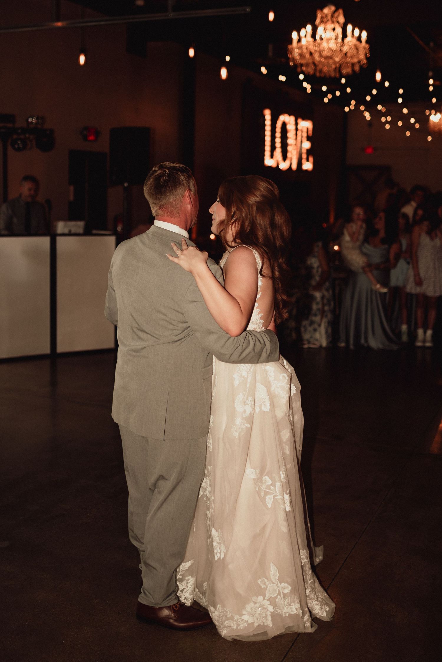 A couple in wedding attire shares a romantic dance on a dimly lit dance floor under twinkling lights and a chandelier.