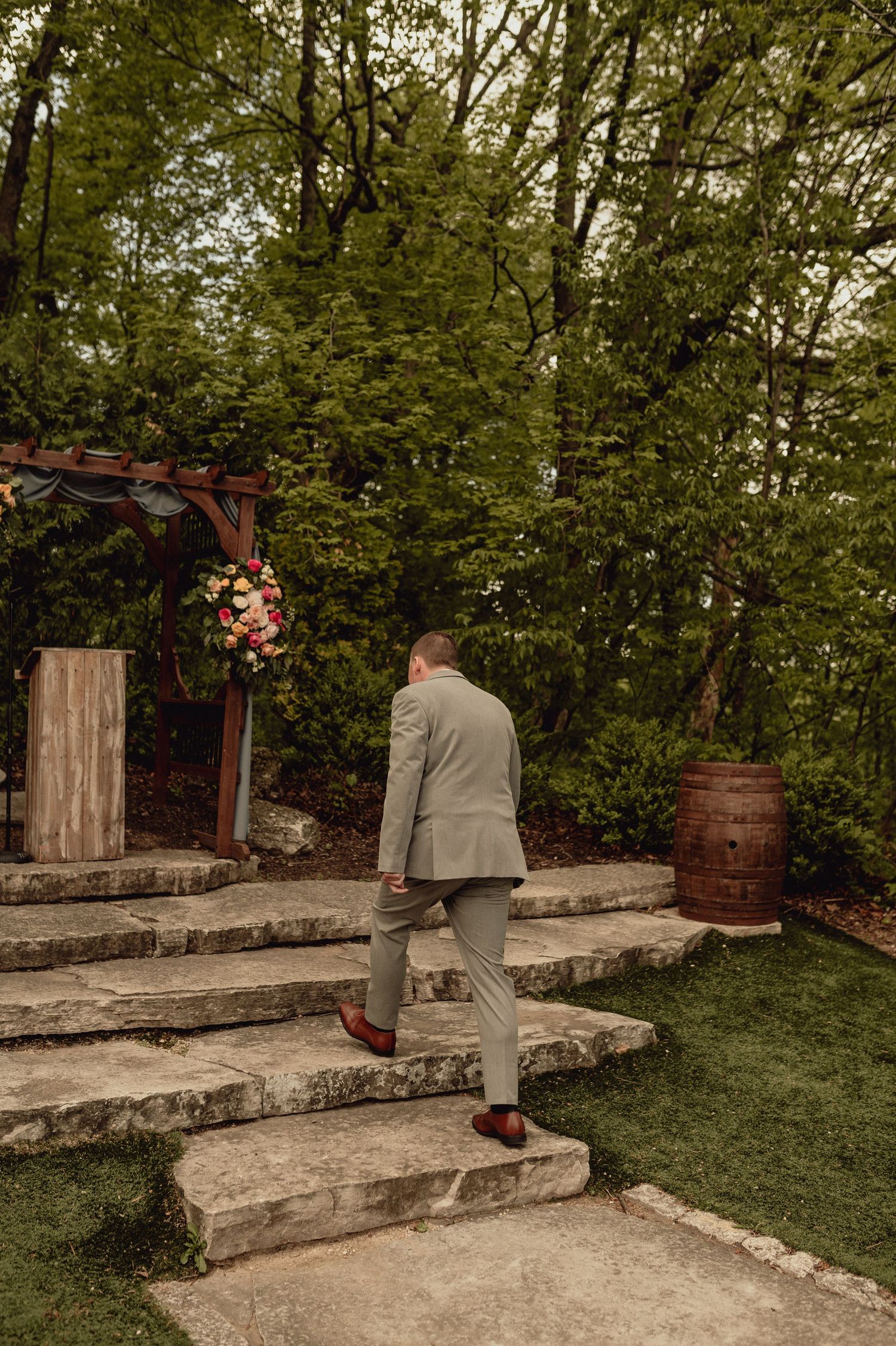 Person in gray suit walks down stone garden steps surrounded by lush greenery and wooden pergola elements.