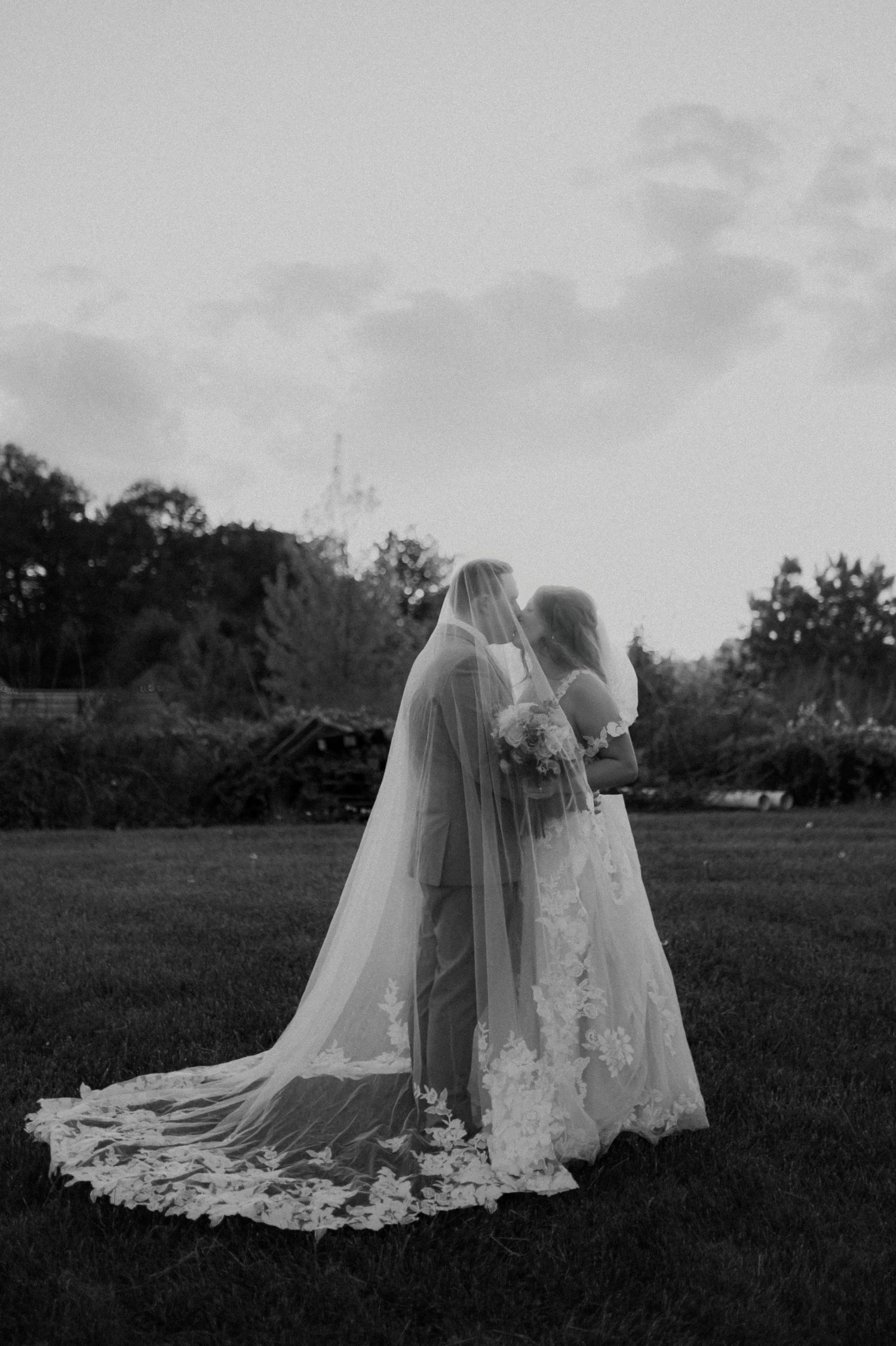 Two figures in flowing white wedding dresses embrace in a misty field at dusk creating a romantic silhouette.