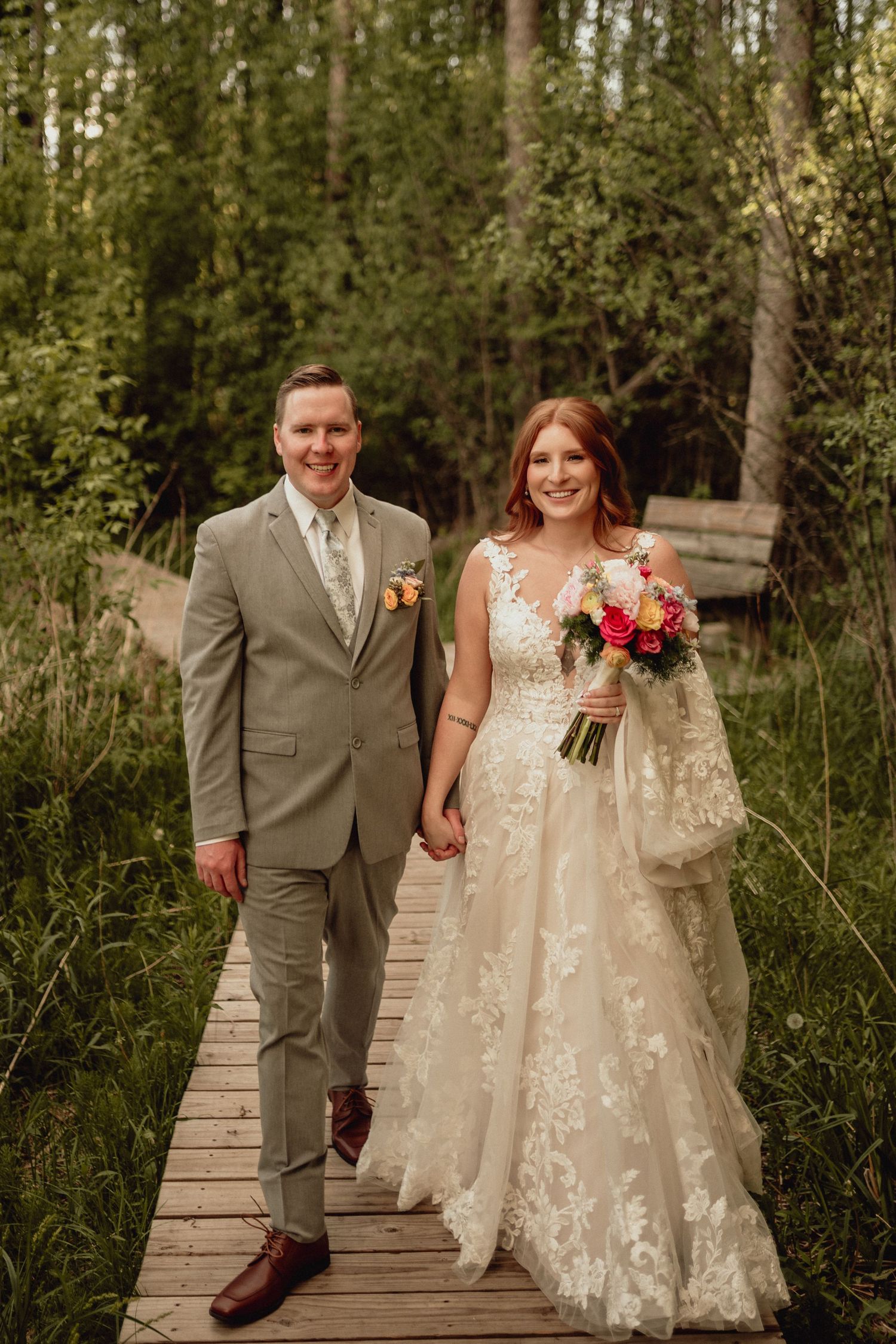 Newlywed couple walks hand in hand on a wooden boardwalk path through lush green forest scenery.