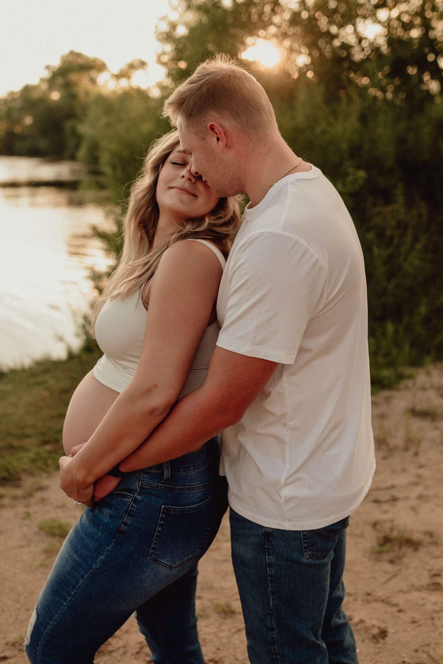 Golden hour maternity photos of couple in jeans sharing intimate moment by lake.
