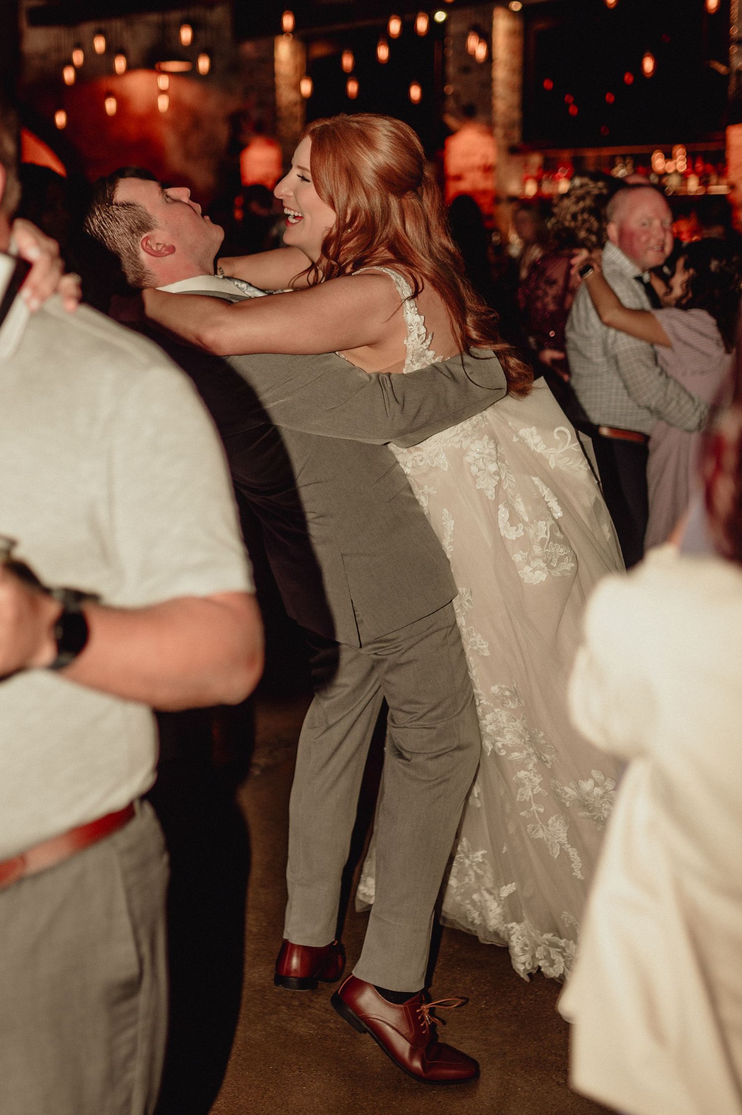 A couple performs a dip dance move at a dimly lit wedding reception with other guests in the background.
