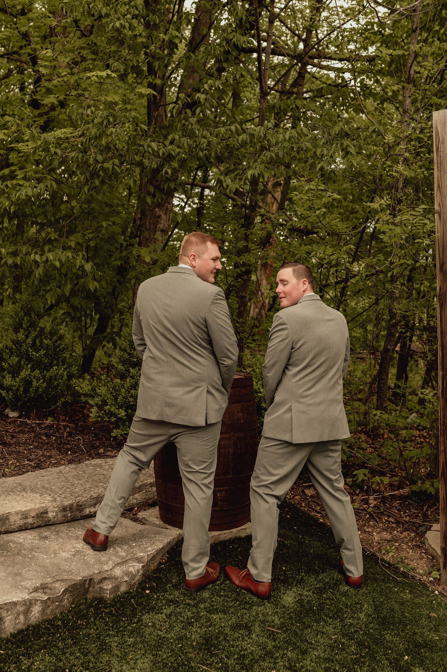 Two men in gray suits share a lighthearted moment on a stone wall surrounded by lush greenery at an outdoor event.