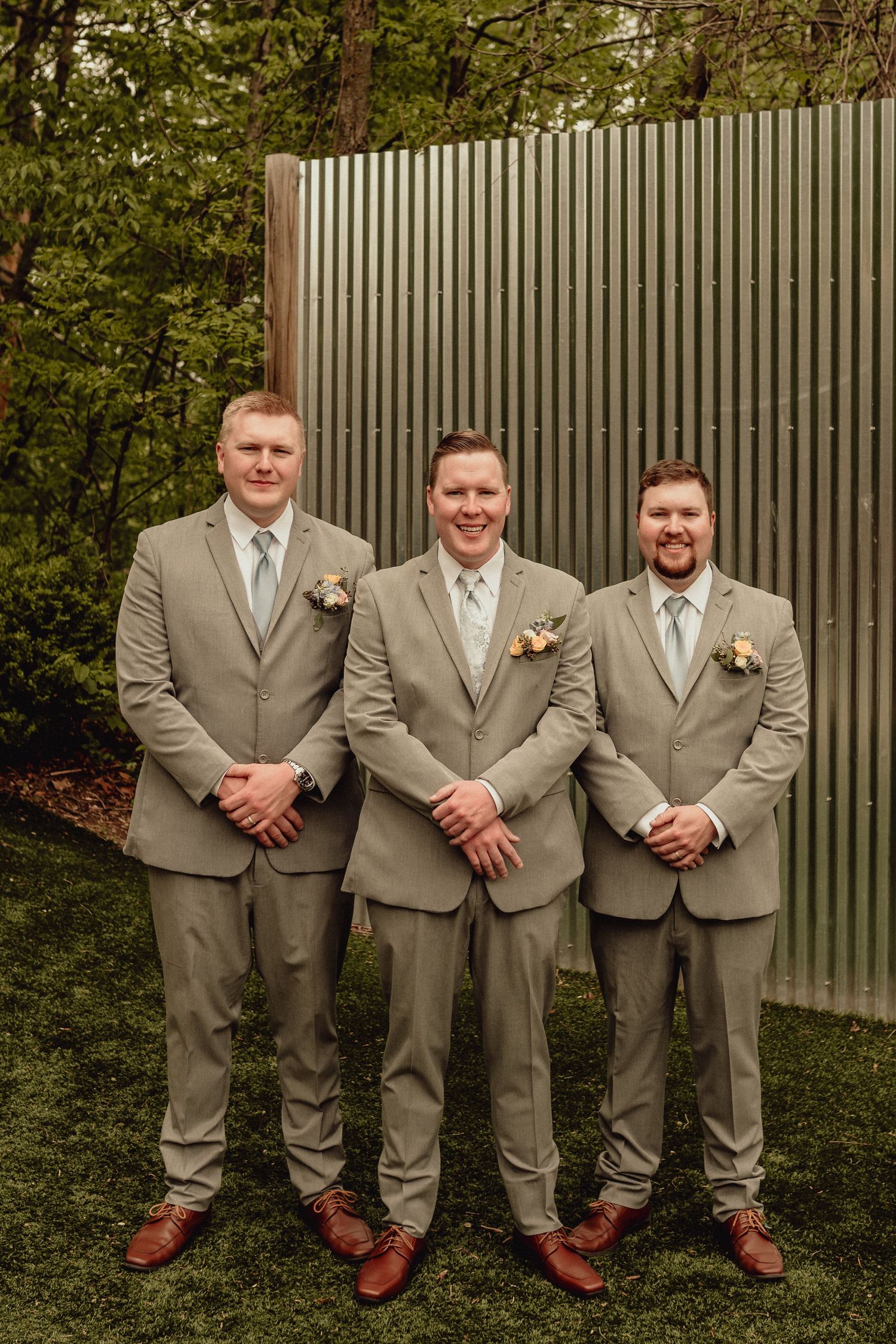 Groomsmen in matching tan suits and brown shoes pose together against a corrugated metal wall and greenery backdrop.