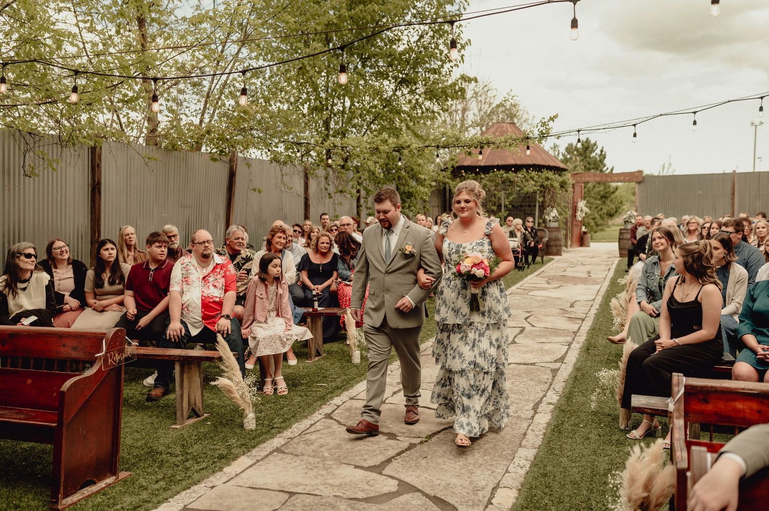 String lights illuminate an outdoor wedding venue with guests mingling on a grassy lawn during golden hour.