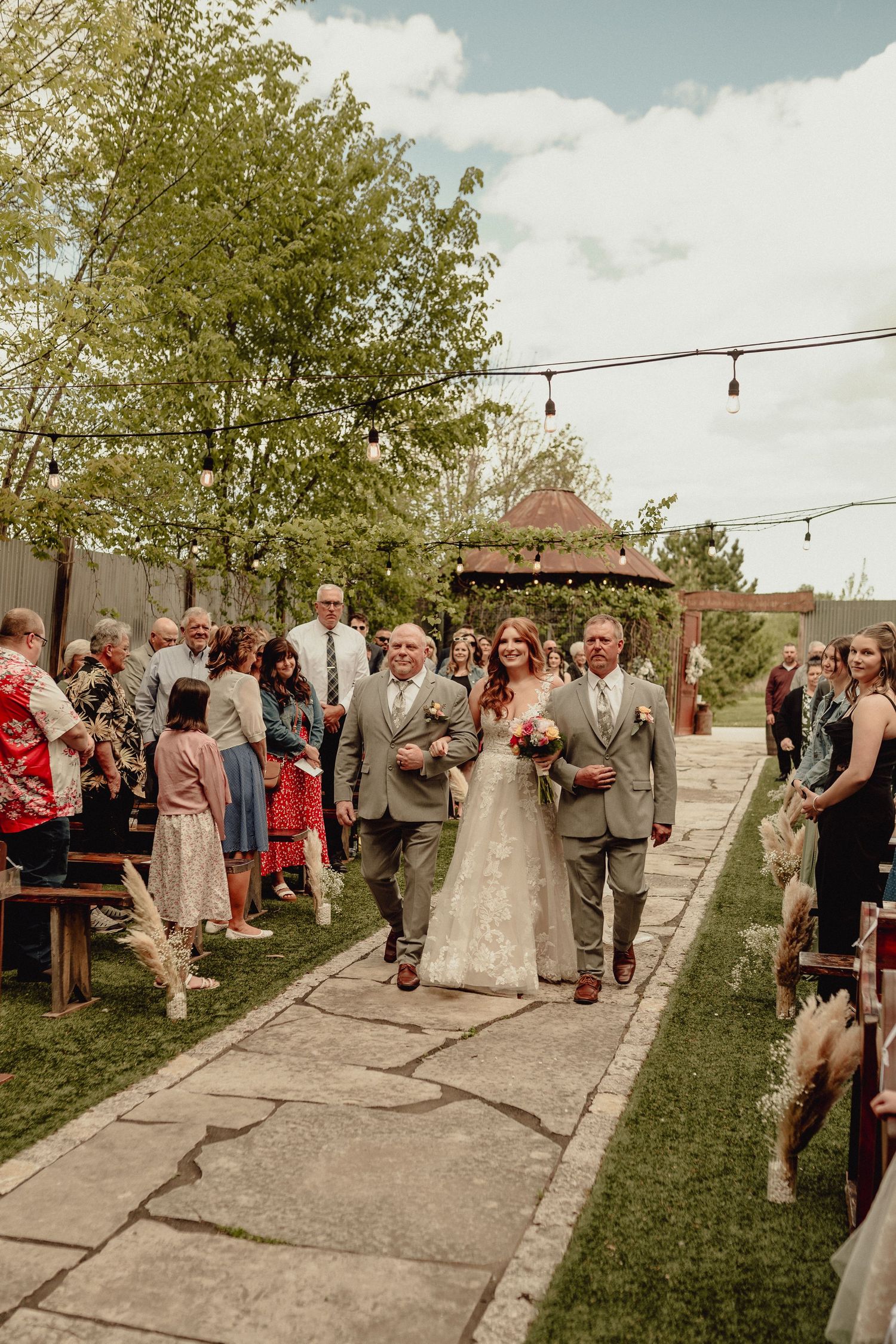 Outdoor wedding ceremony guests gathered along a stone path under string lights and trees on a sunny day.