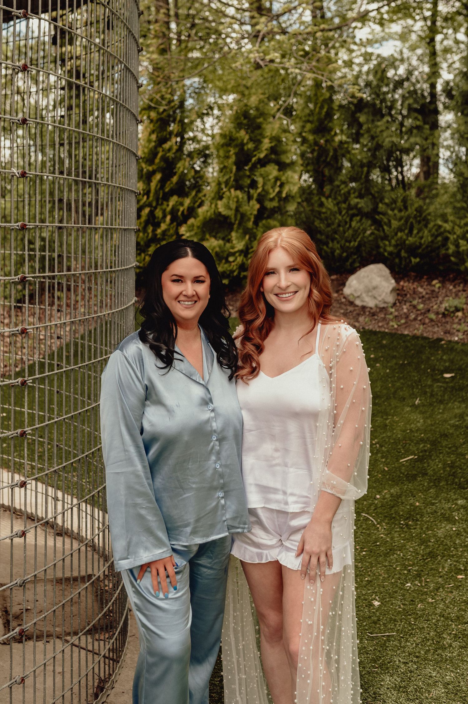Two friends in casual attire pose playfully against an outdoor metal fence backdrop.
