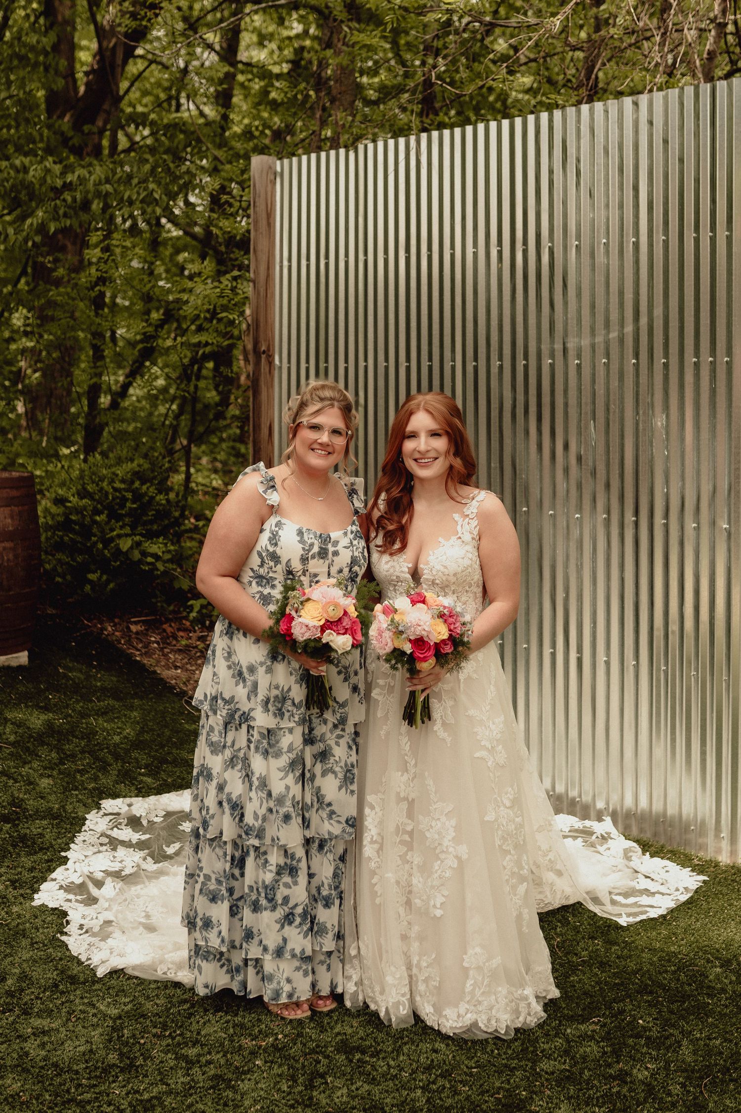 Two people in elegant dresses pose together outdoors against a white fence holding colorful floral bouquets.