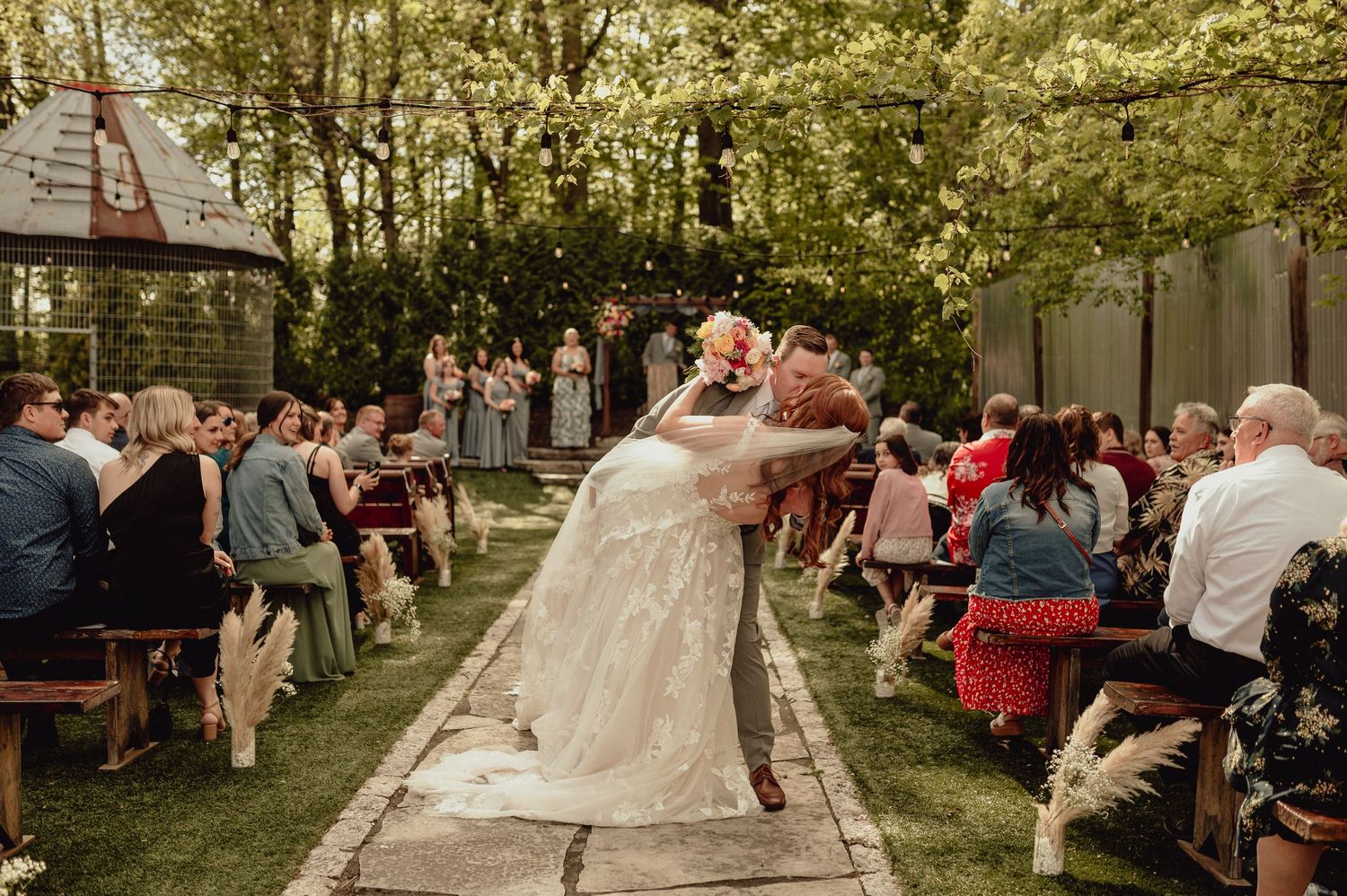 Wedding guests dance and celebrate outdoors under a canopy of trees at a rustic reception venue.