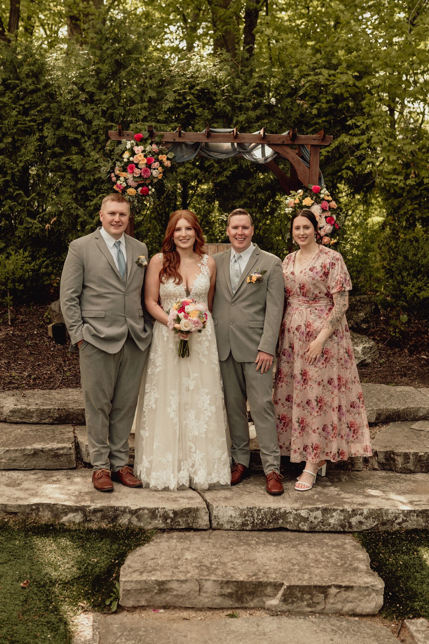 A wedding party stands on stone steps under a floral arch in a garden setting, wearing gray suits and floral dresses.