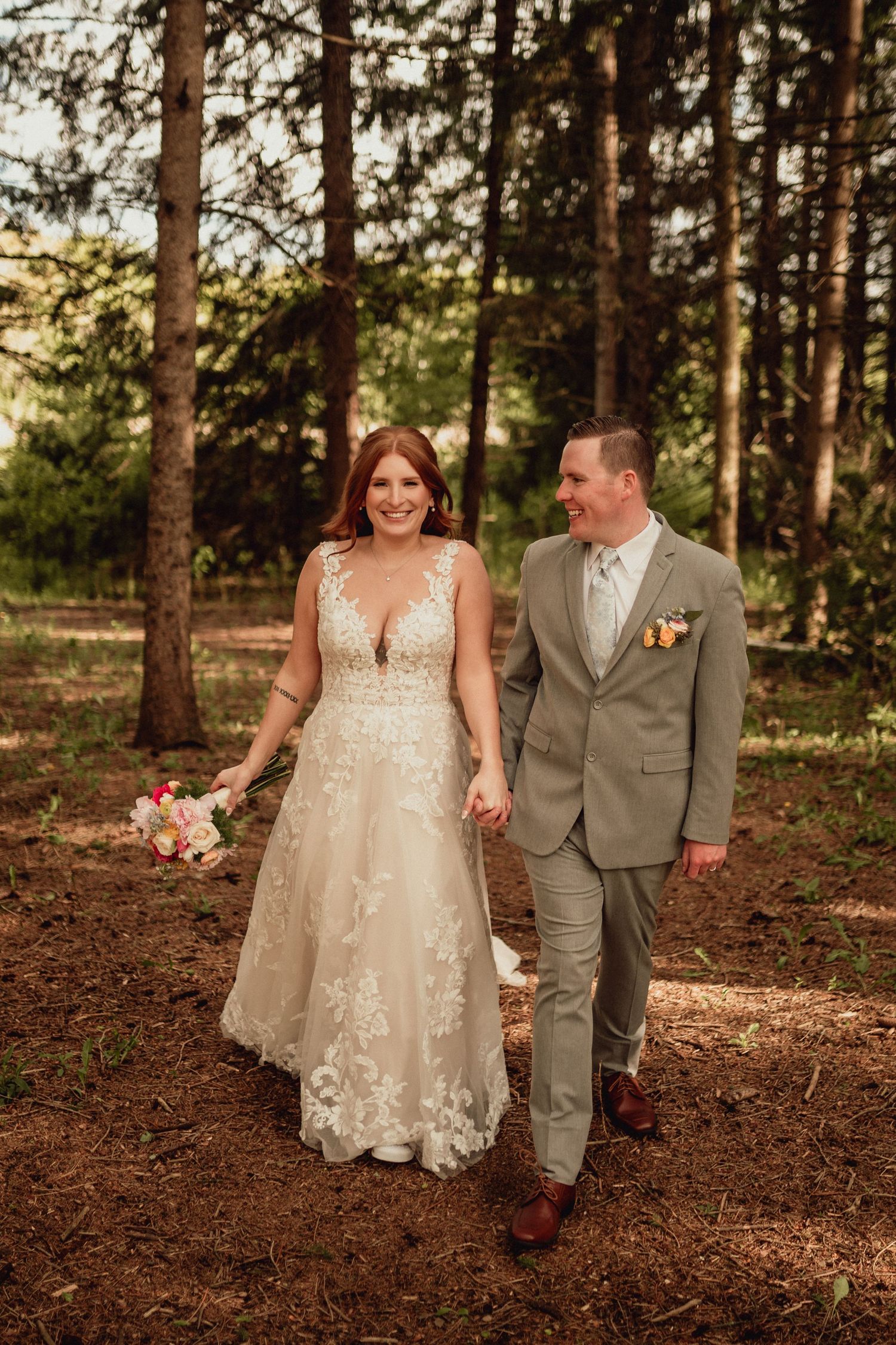 A couple walks hand in hand through a pine forest during their wedding, with the bride wearing a lace gown and groom in gray.