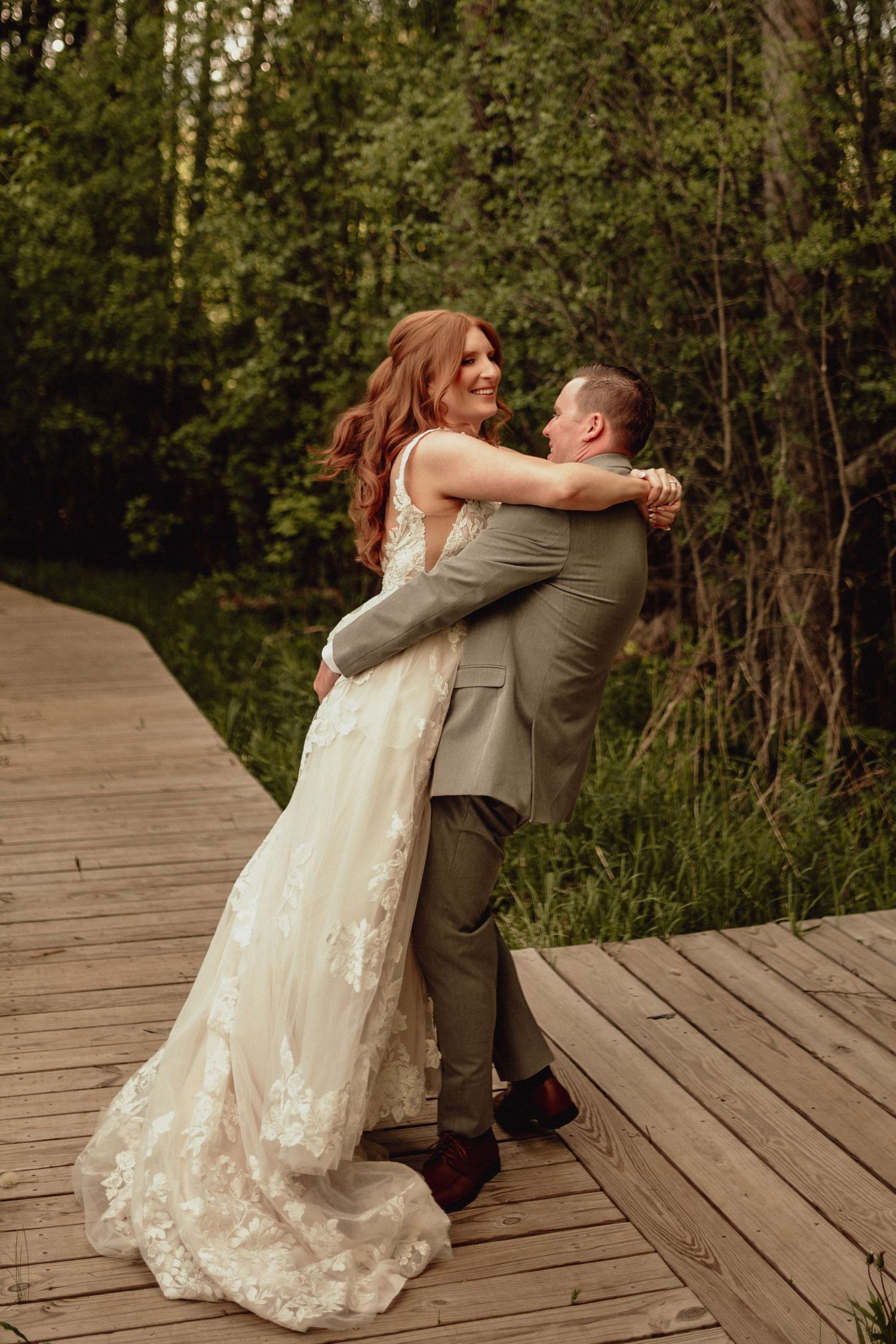 A couple in wedding attire share a romantic moment on a wooden boardwalk surrounded by greenery.