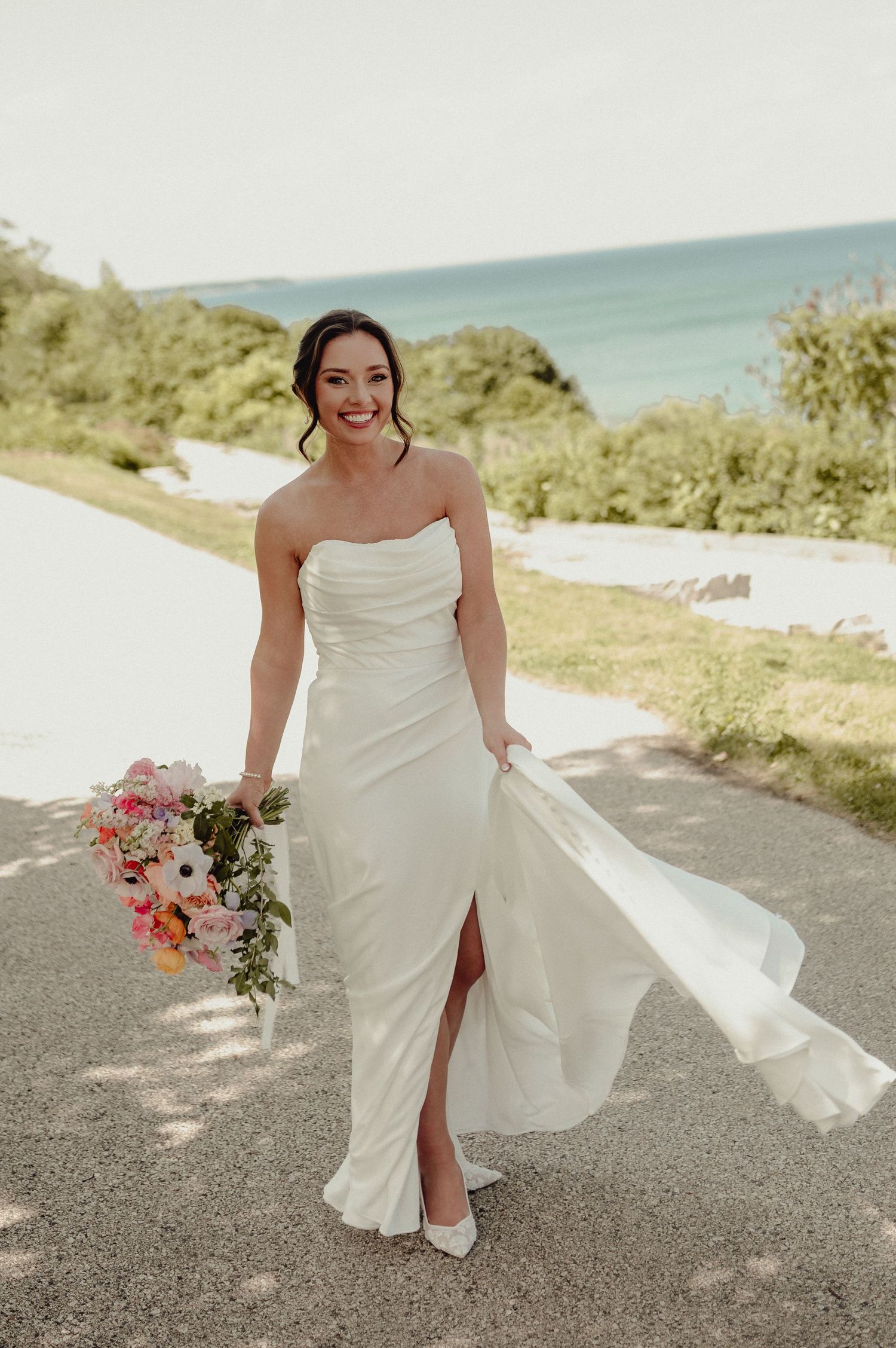 Bride in flowing white dress holding bouquet while walking along coastal road.