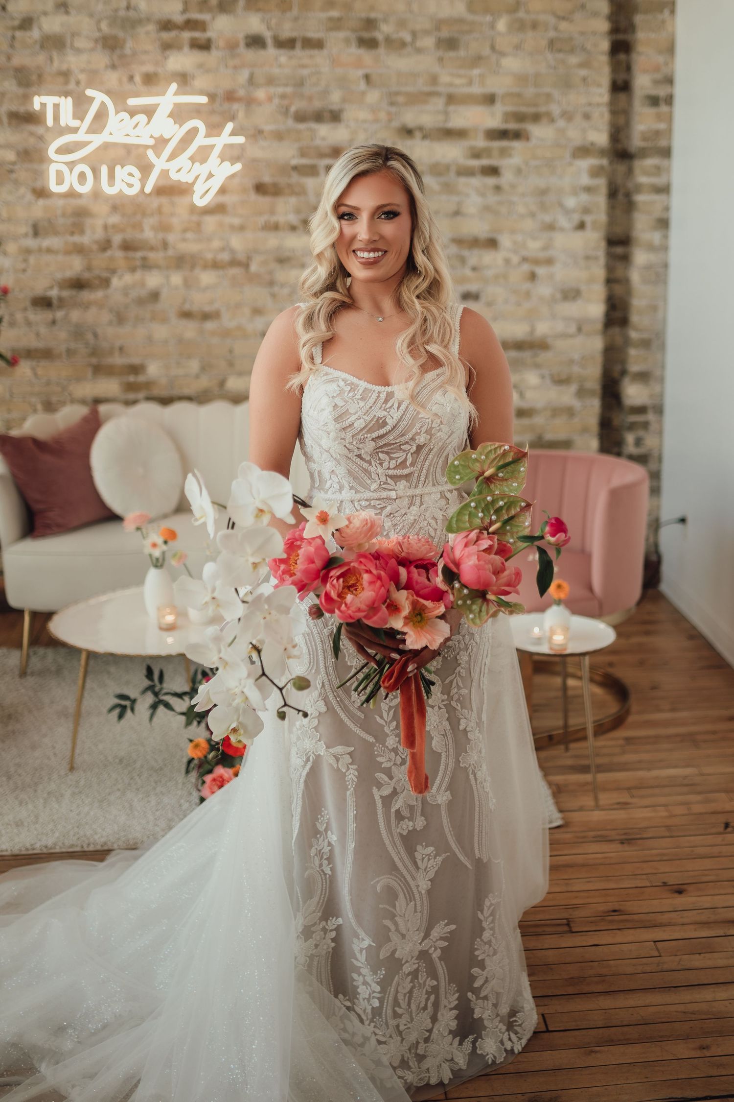 Bride in detailed lace gown poses with elegant floral arrangements in rustic brick venue.