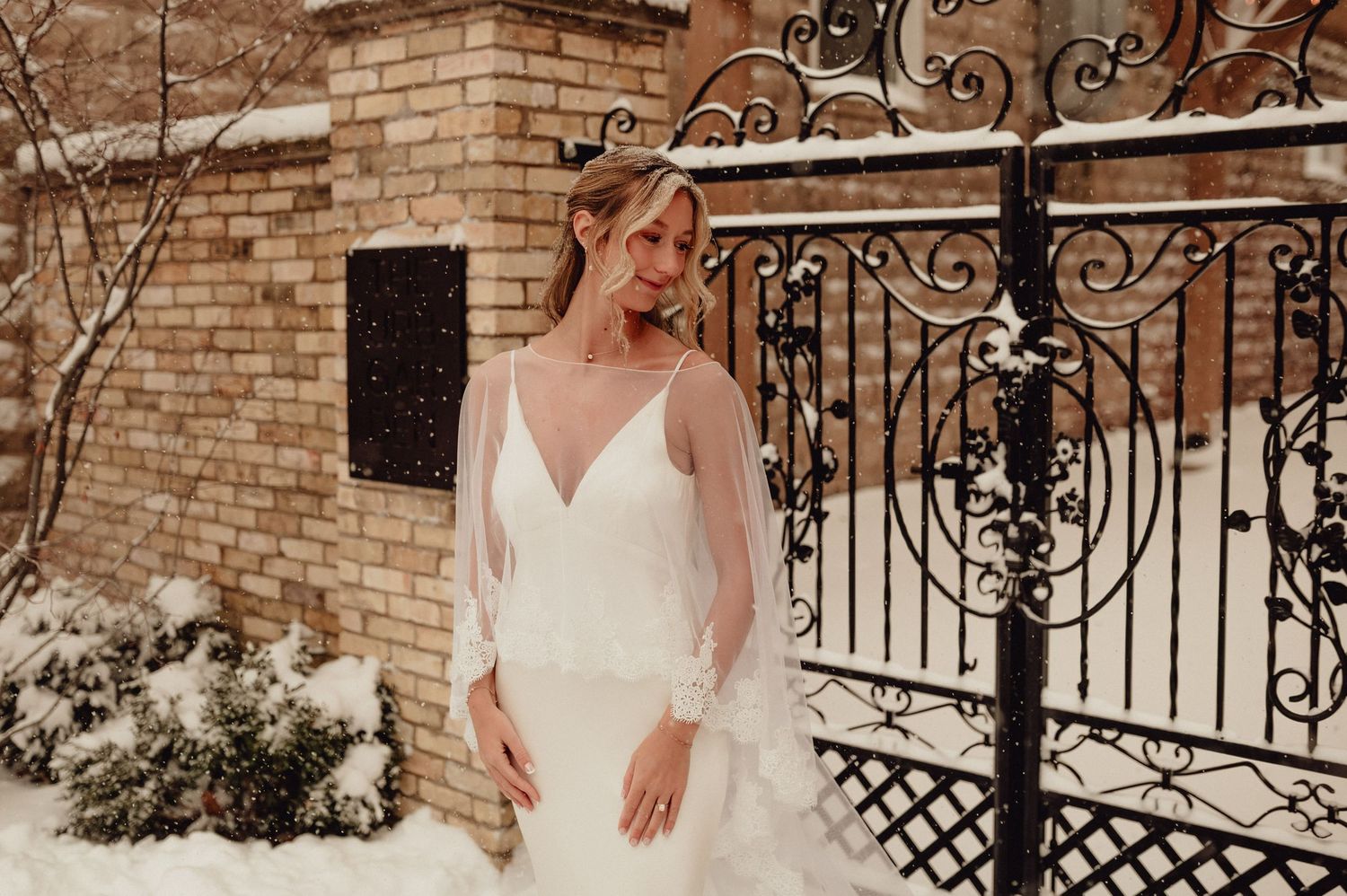 Bride in elegant white wedding dress poses gracefully by ornate wrought iron gate and brick wall.