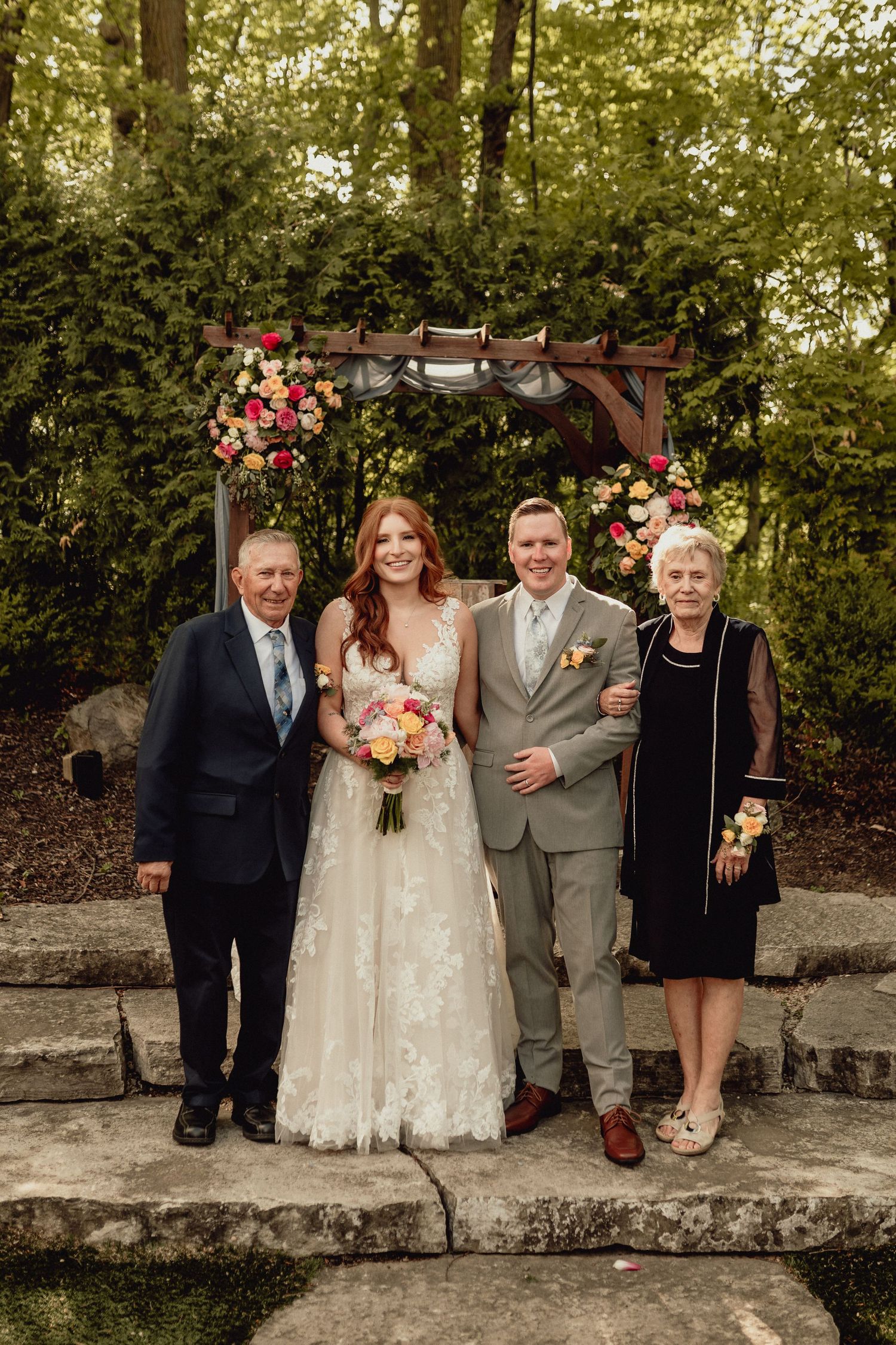 Wedding party poses together on stone steps in an outdoor garden setting with lush greenery in the background.