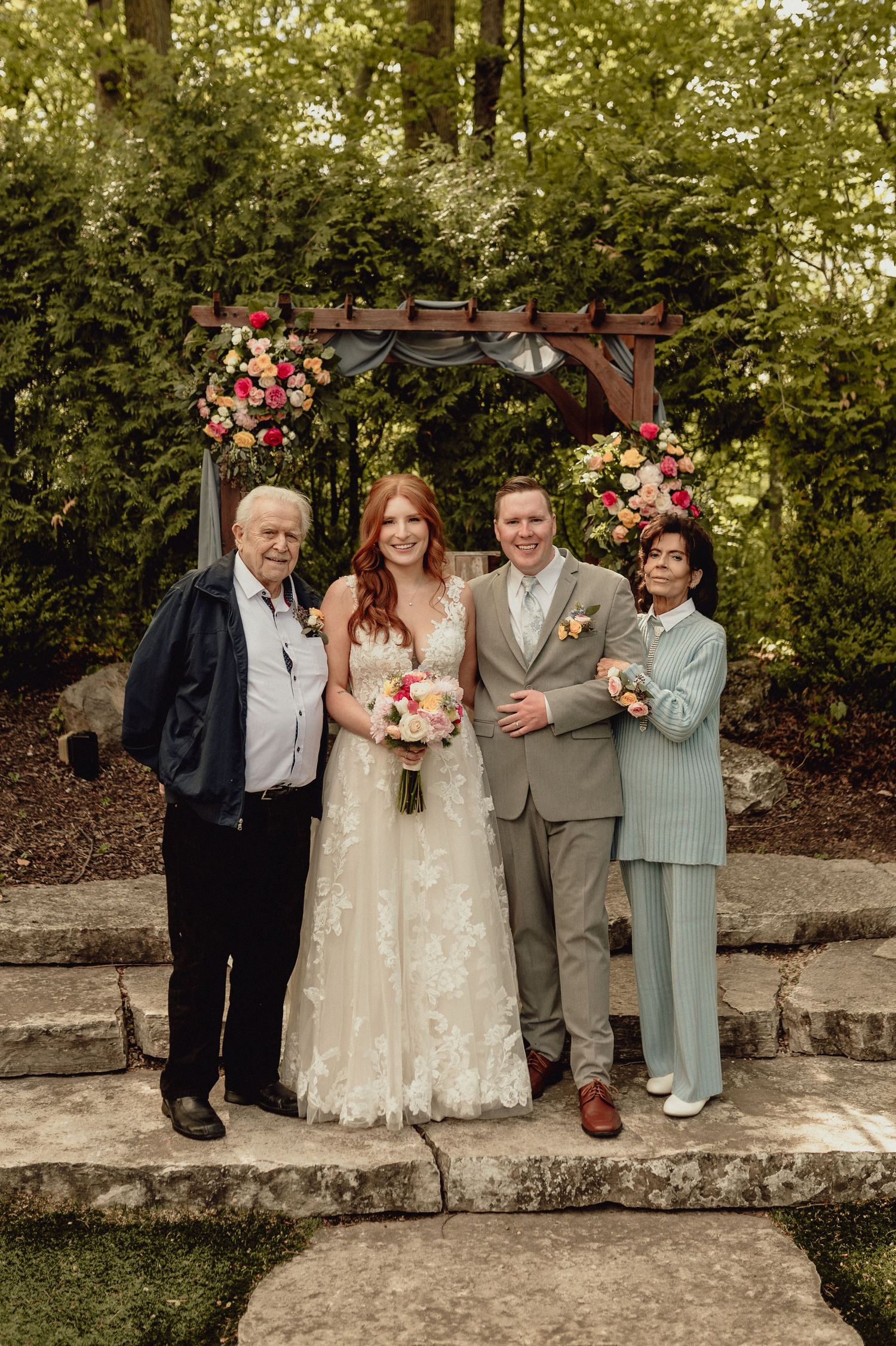 A wedding party poses under a floral arch in an outdoor garden setting with greenery in the background.