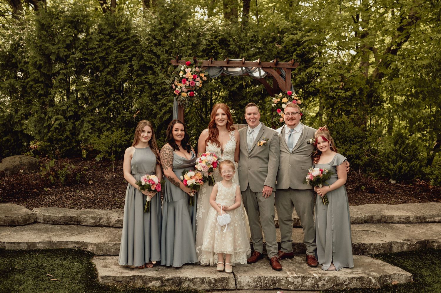 Wedding party in gray and white attire poses together outdoors in a garden setting during a summer celebration.
