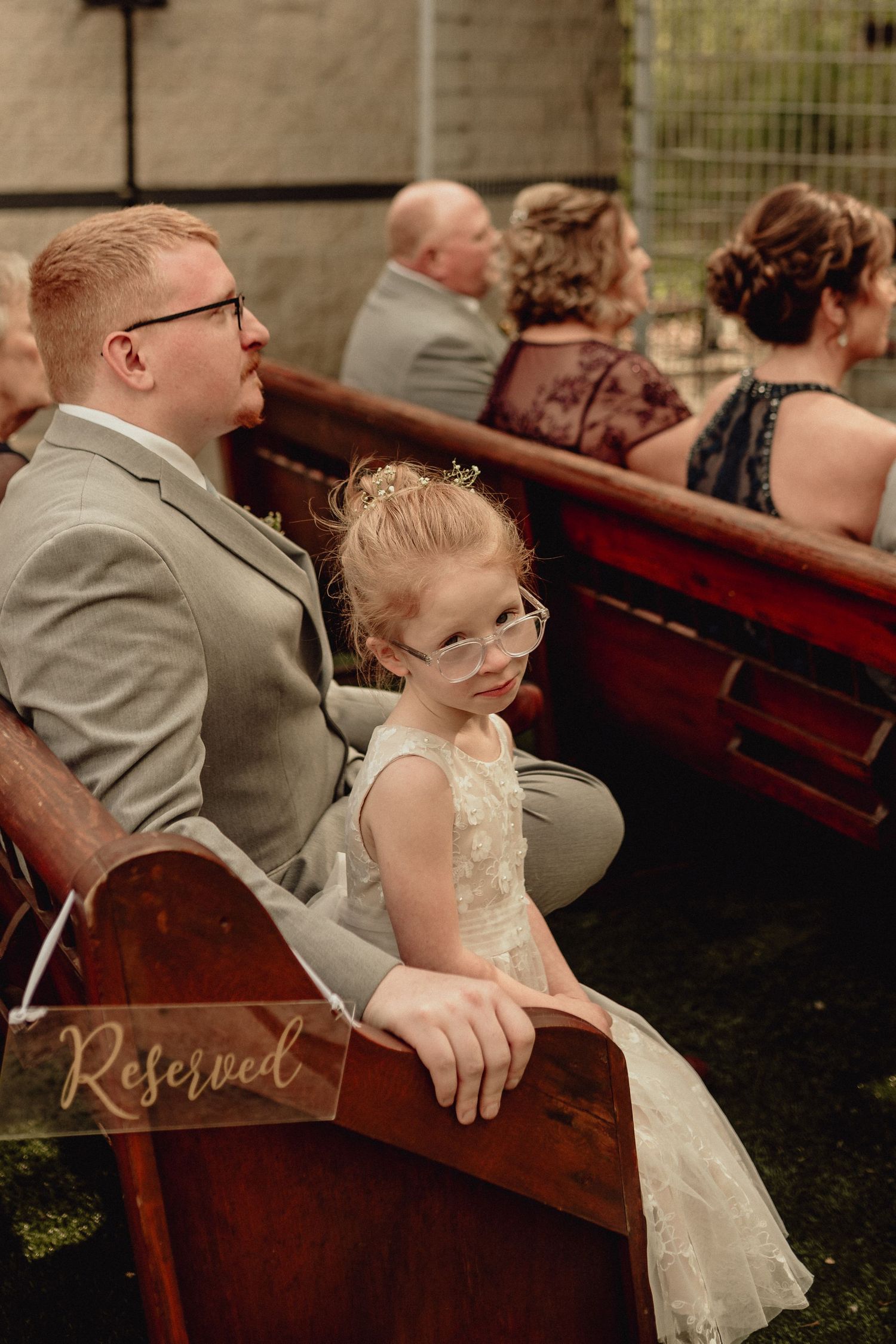 A young girl in a white dress and glasses sits next to an adult in a gray suit on wooden church pews marked 'Reserved'.