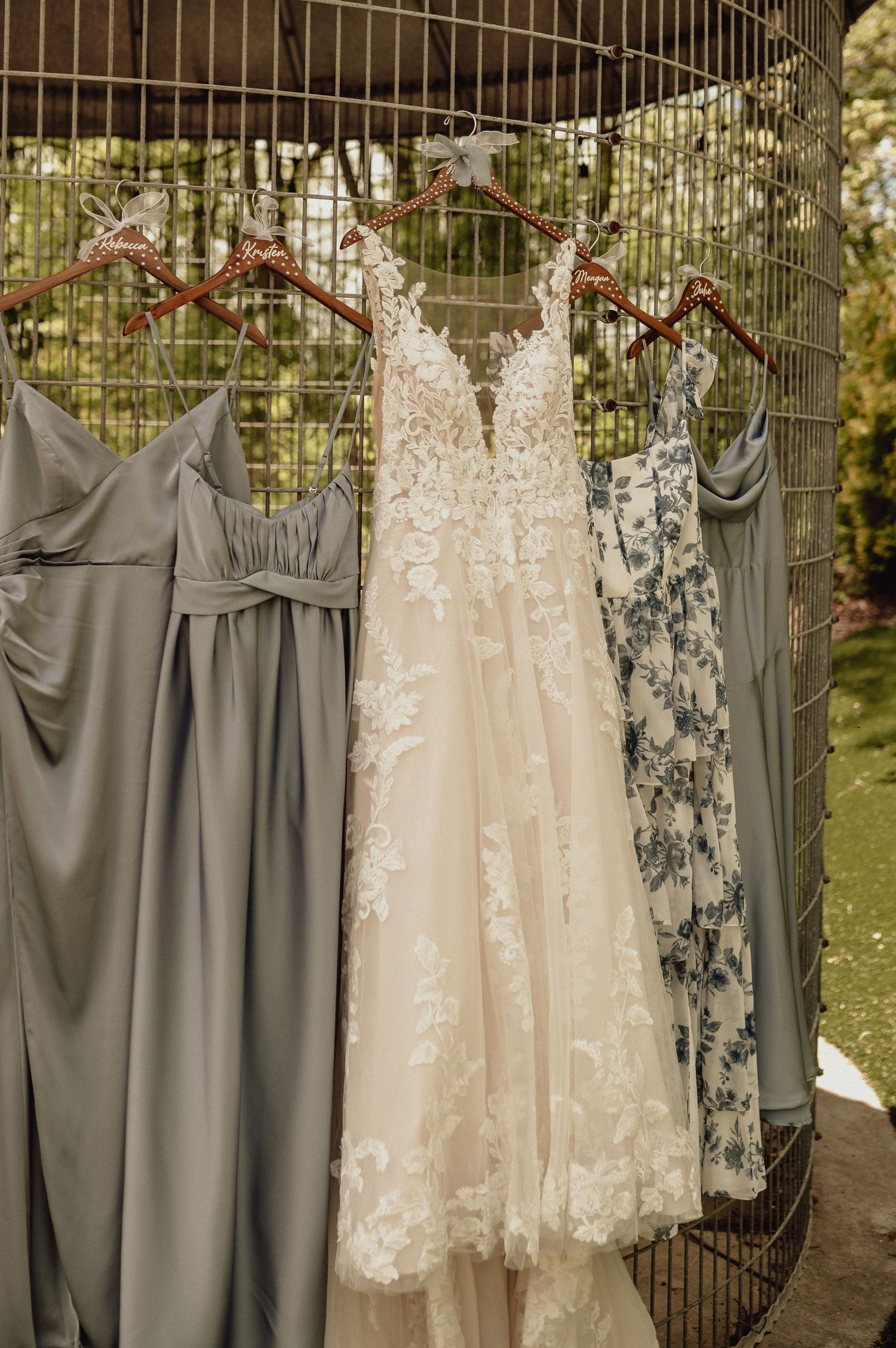 Wedding dresses and bridesmaid gowns in gray and cream colors hang on display outdoors against an iron fence.