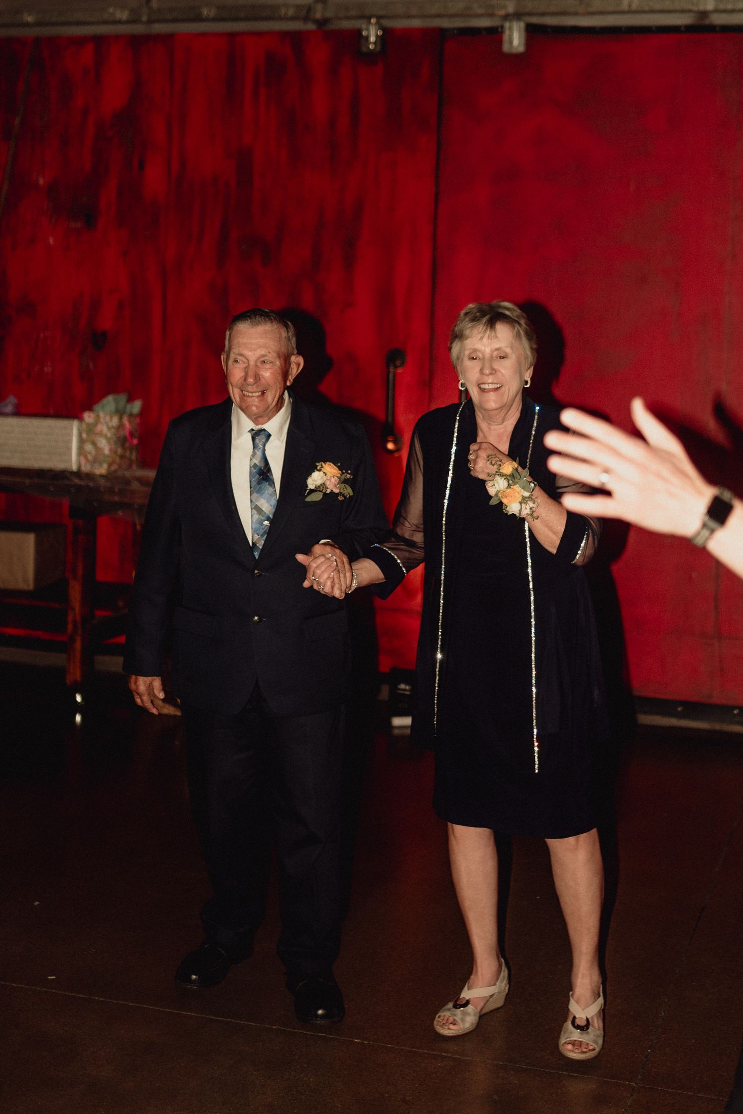 An elderly couple dances together on a dark dance floor with red curtains in the background.