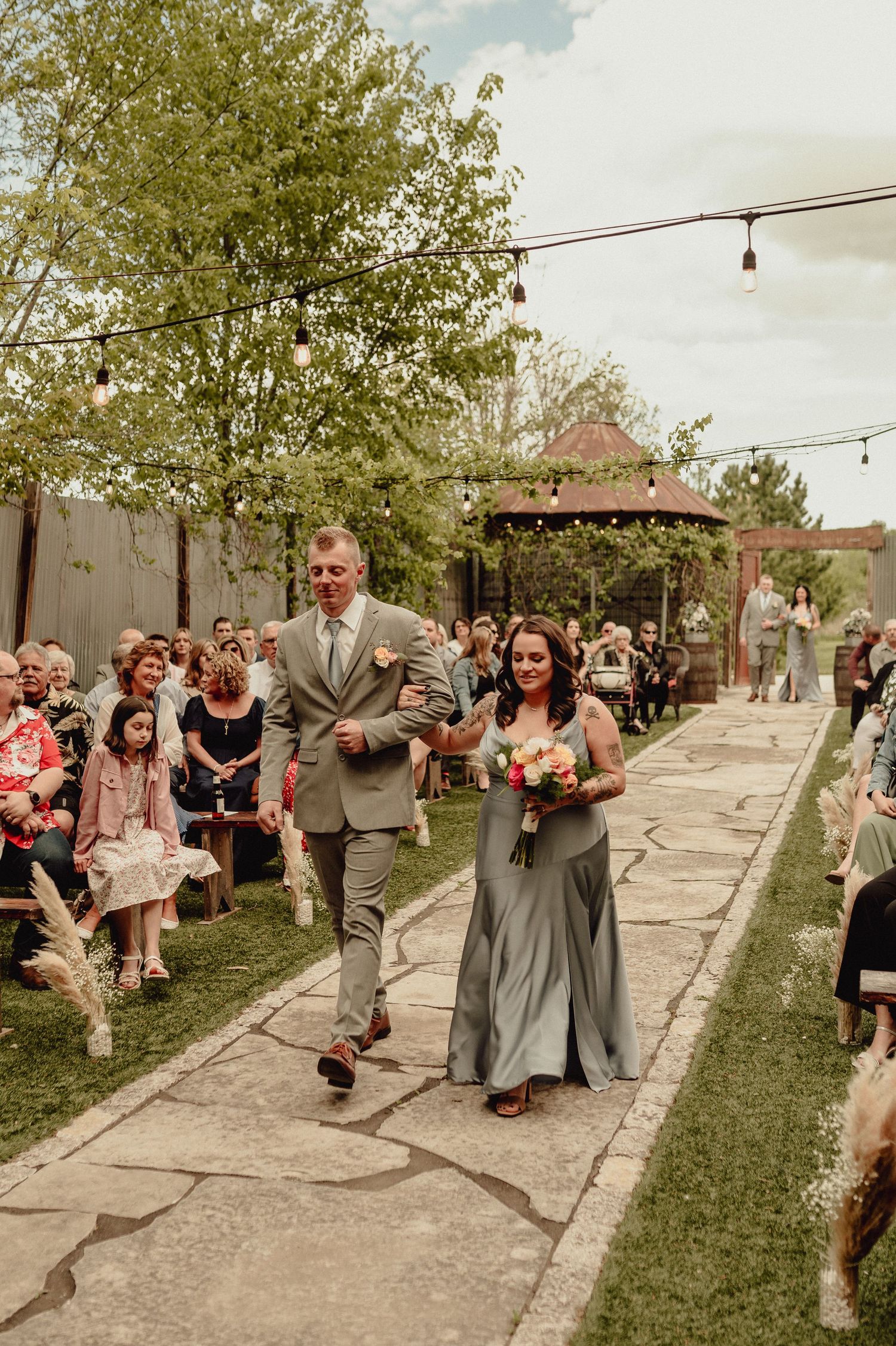 Wedding party dances in a line on a stone pathway surrounded by grass and guests during an outdoor celebration.