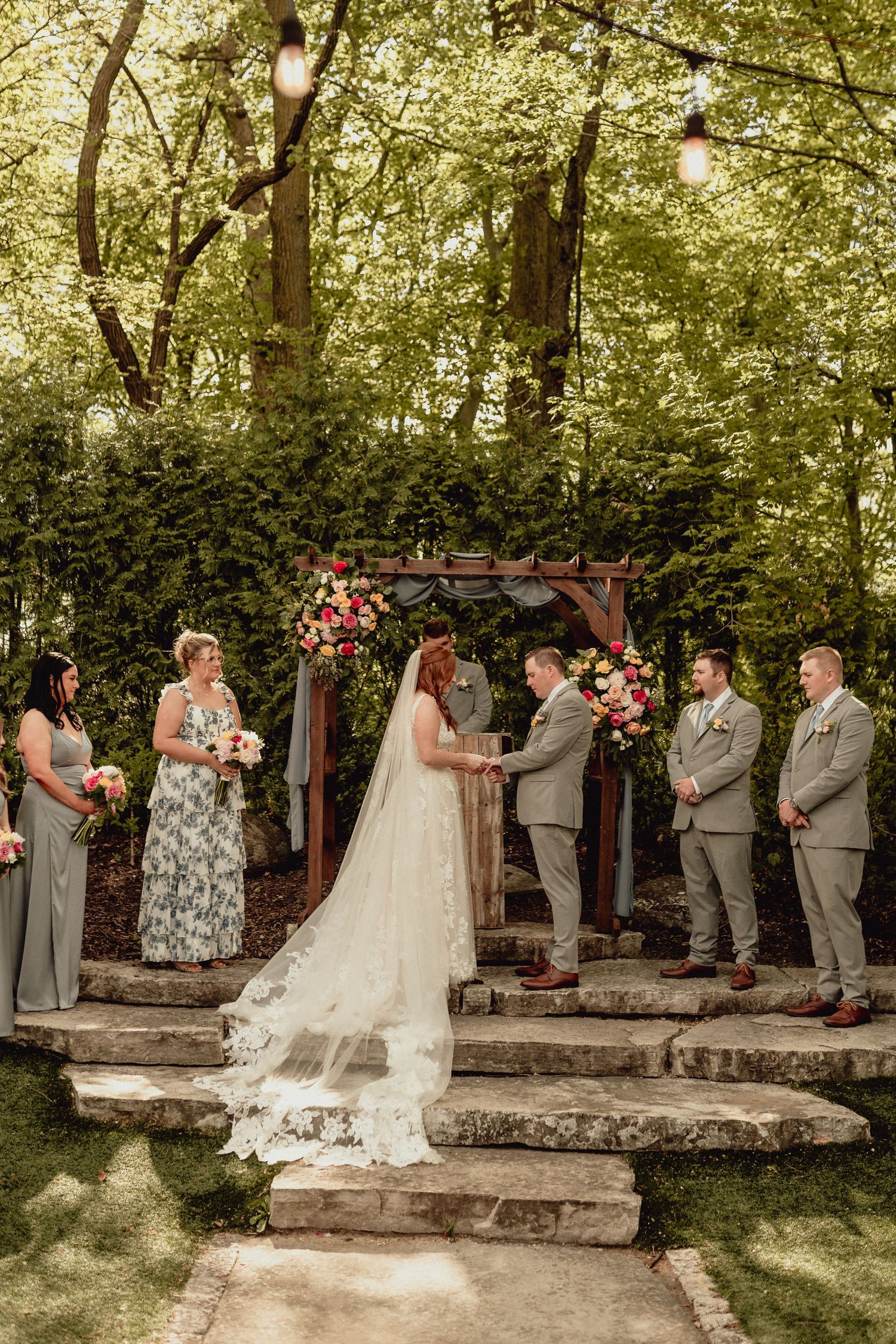 Wedding party stands in line during outdoor garden ceremony with stone steps and greenery in the background.