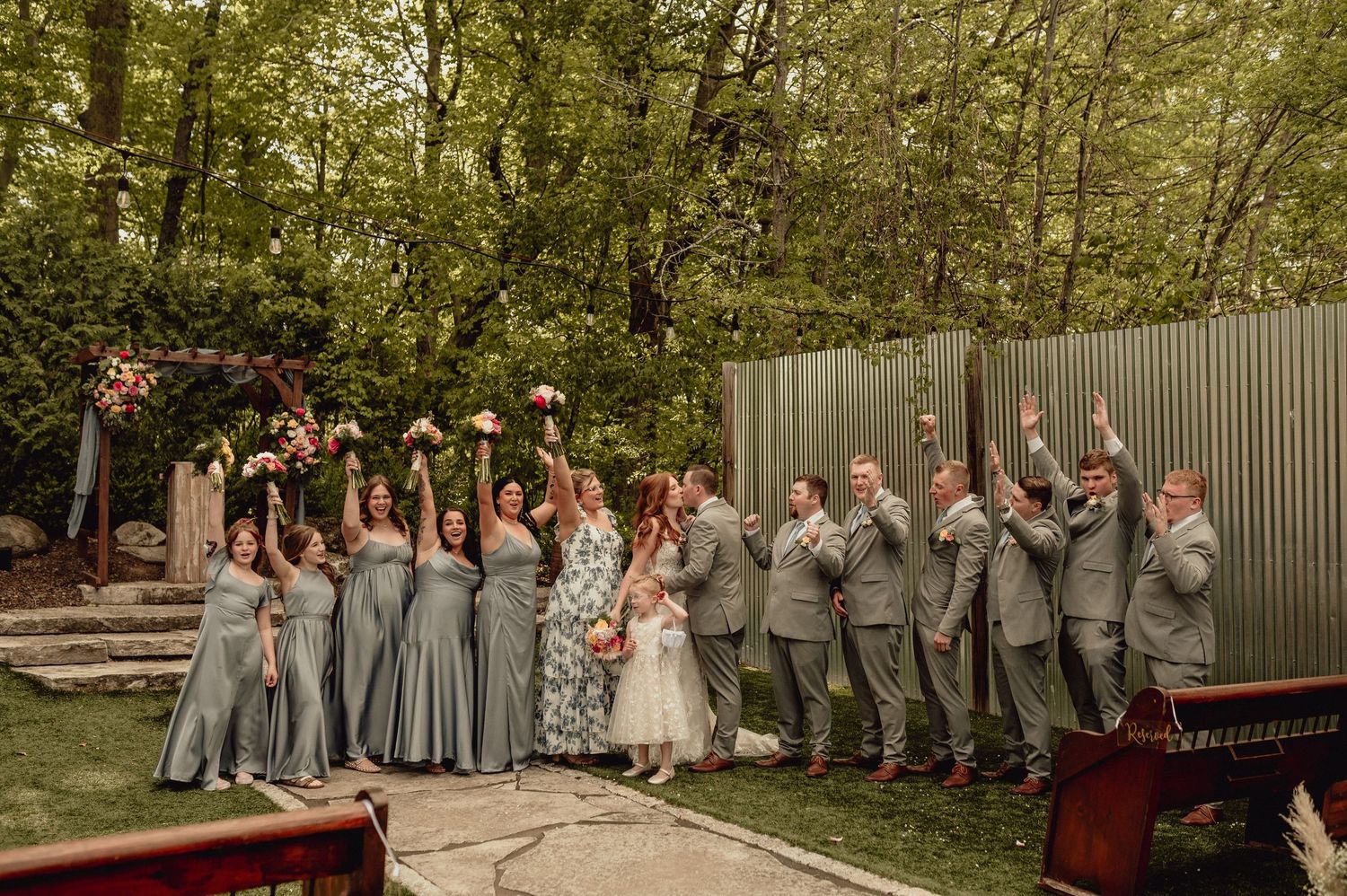 Wedding party in gray attire jumps joyfully for a group photo in a wooded outdoor setting.