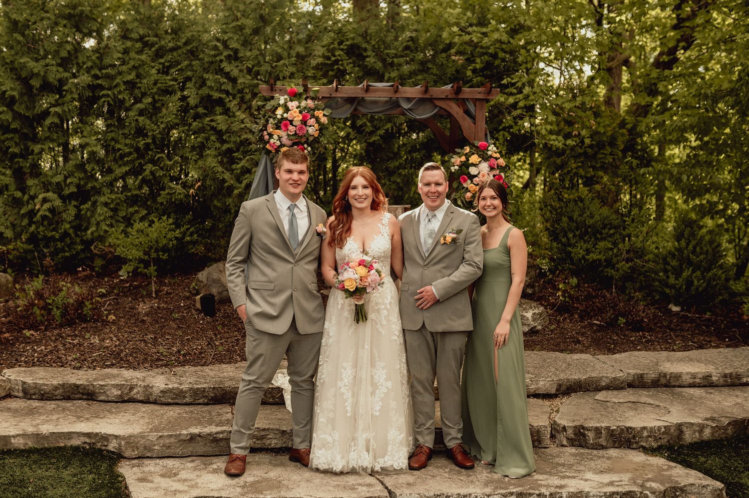 A wedding party poses together on stone steps surrounded by lush greenery in an outdoor garden setting.