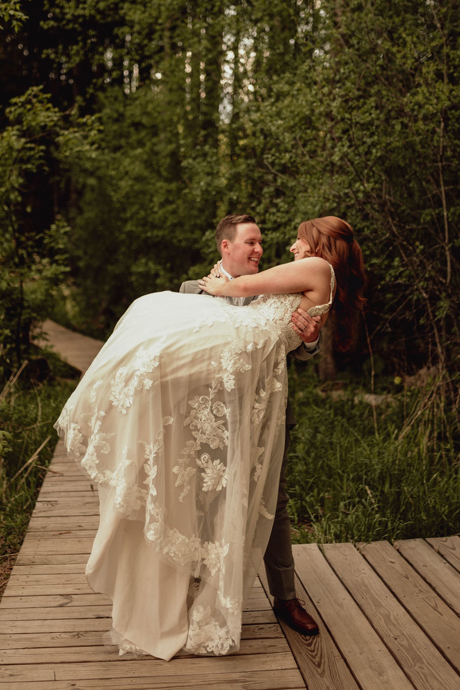 A romantic wedding couple shares a playful dip pose on a wooden boardwalk surrounded by lush forest greenery.