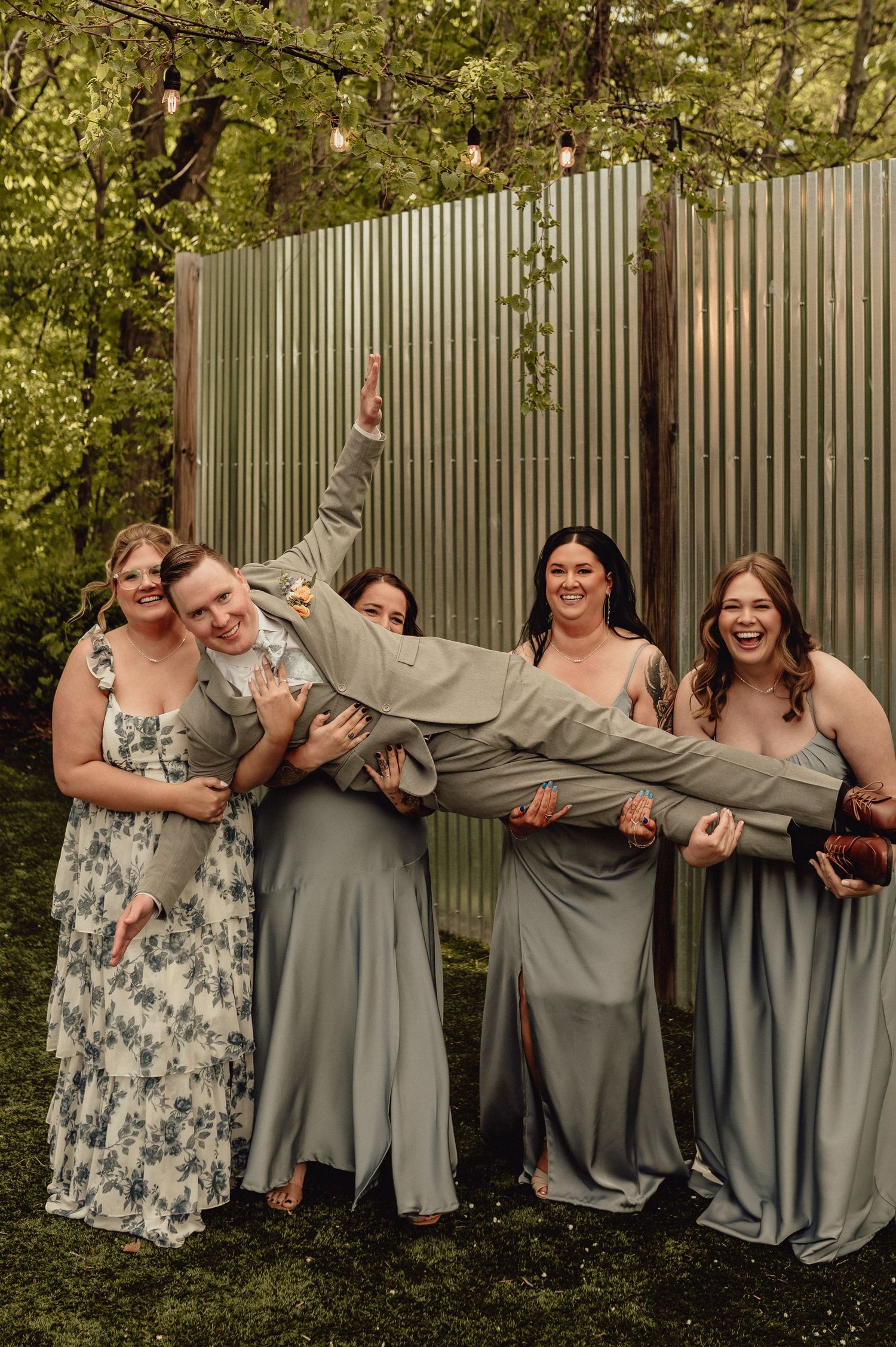 A group in gray and floral dresses poses playfully for a series of candid photos outdoors against a fence.
