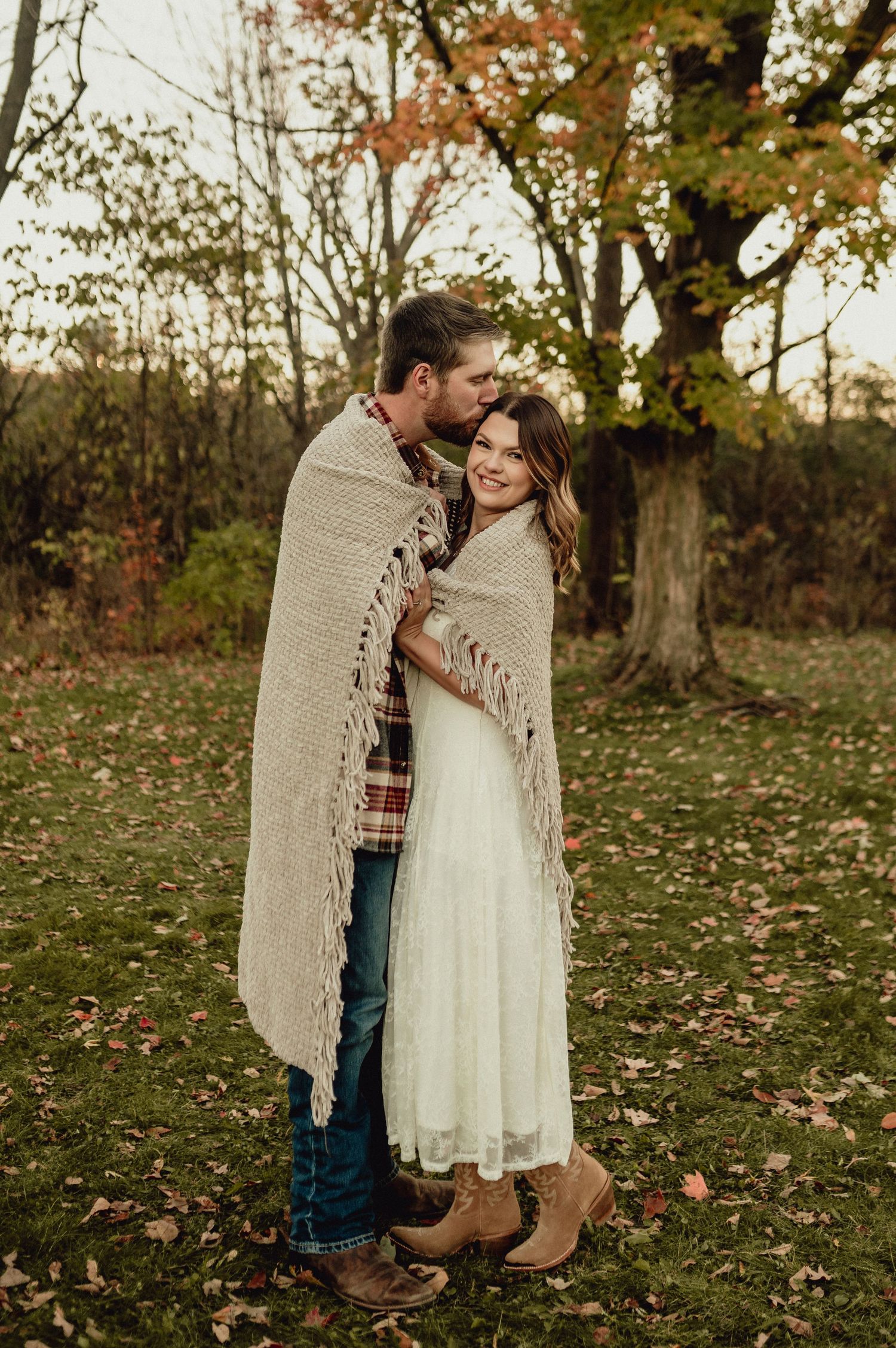 Couple wrapped in beige blanket embracing in autumn forest setting.