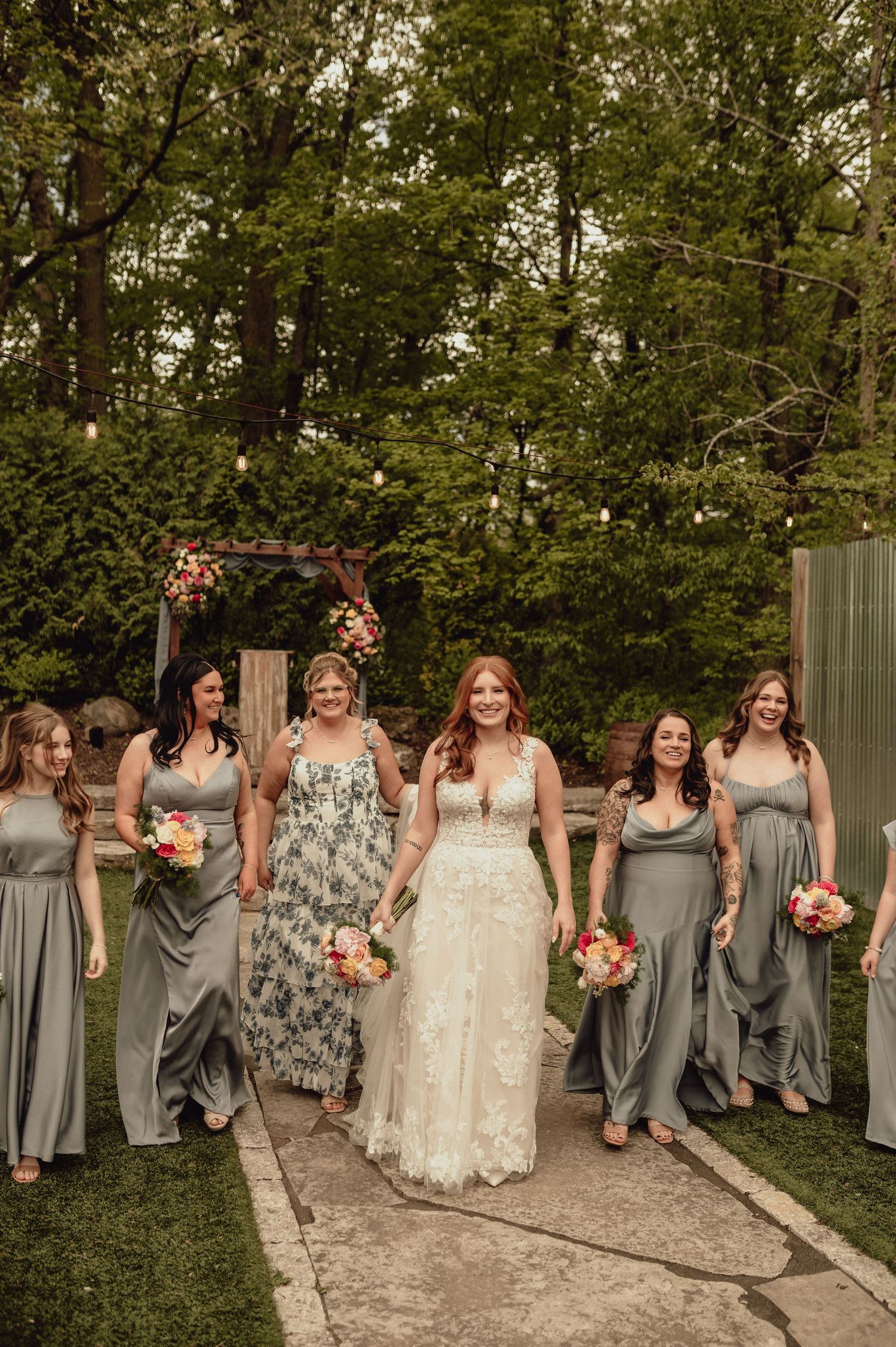 A bridal party in gray bridesmaid dresses and a white wedding gown walk along a garden path under string lights.