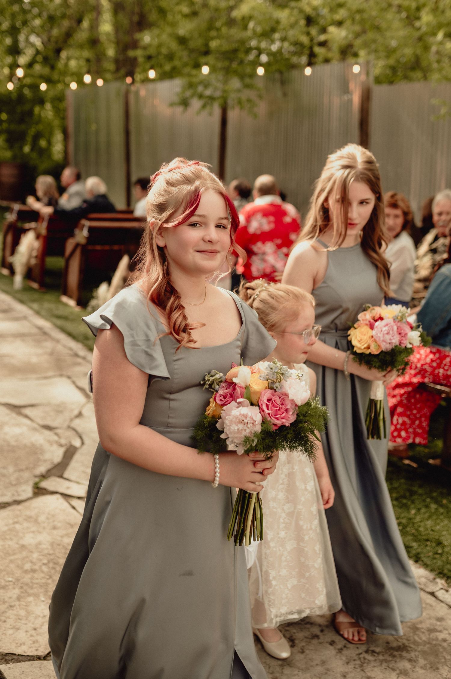 Wedding party members in gray dresses hold pink and white floral bouquets during an outdoor celebration.