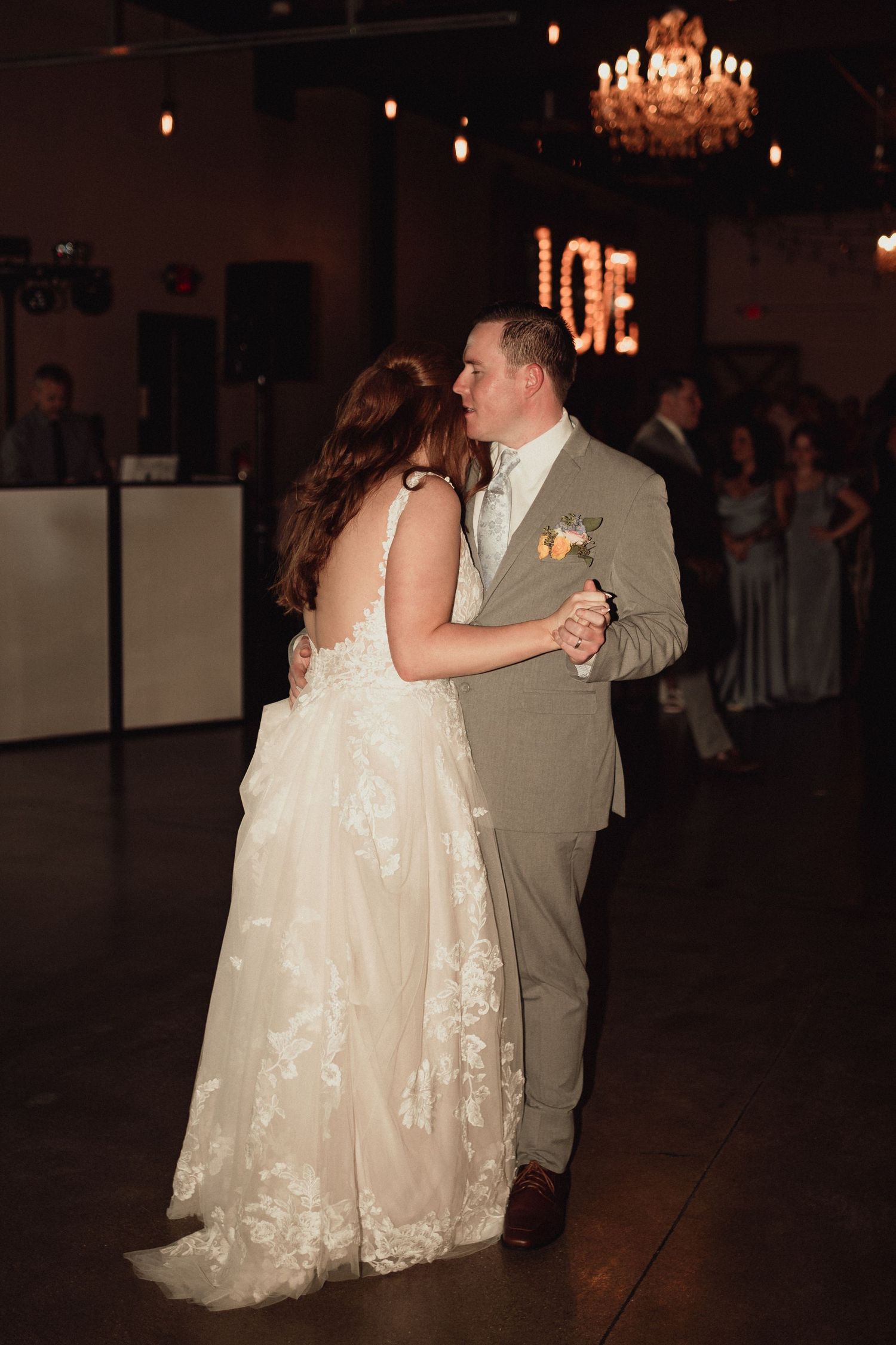 A couple shares their first dance at their wedding reception under a crystal chandelier in a dimly lit ballroom.
