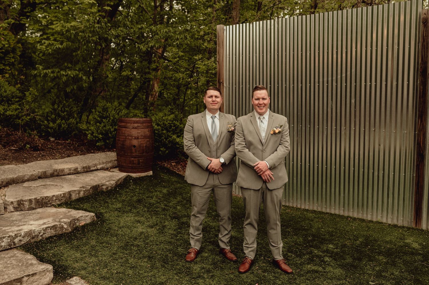 Wedding party members sit casually on stone steps in a garden setting wearing light colored suits.