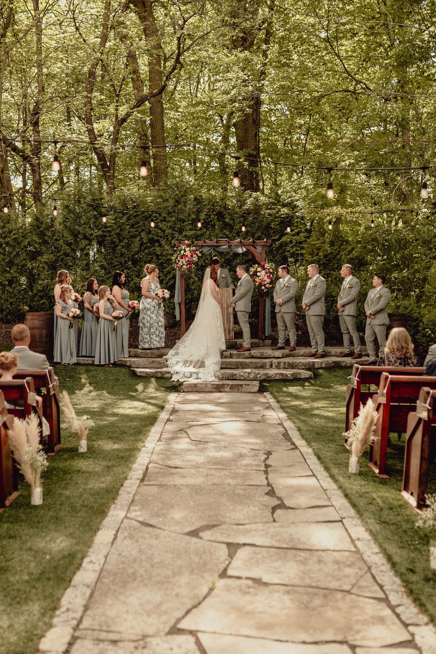 Outdoor wedding ceremony in a wooded setting with stone pathway and wooden pews arranged on either side.